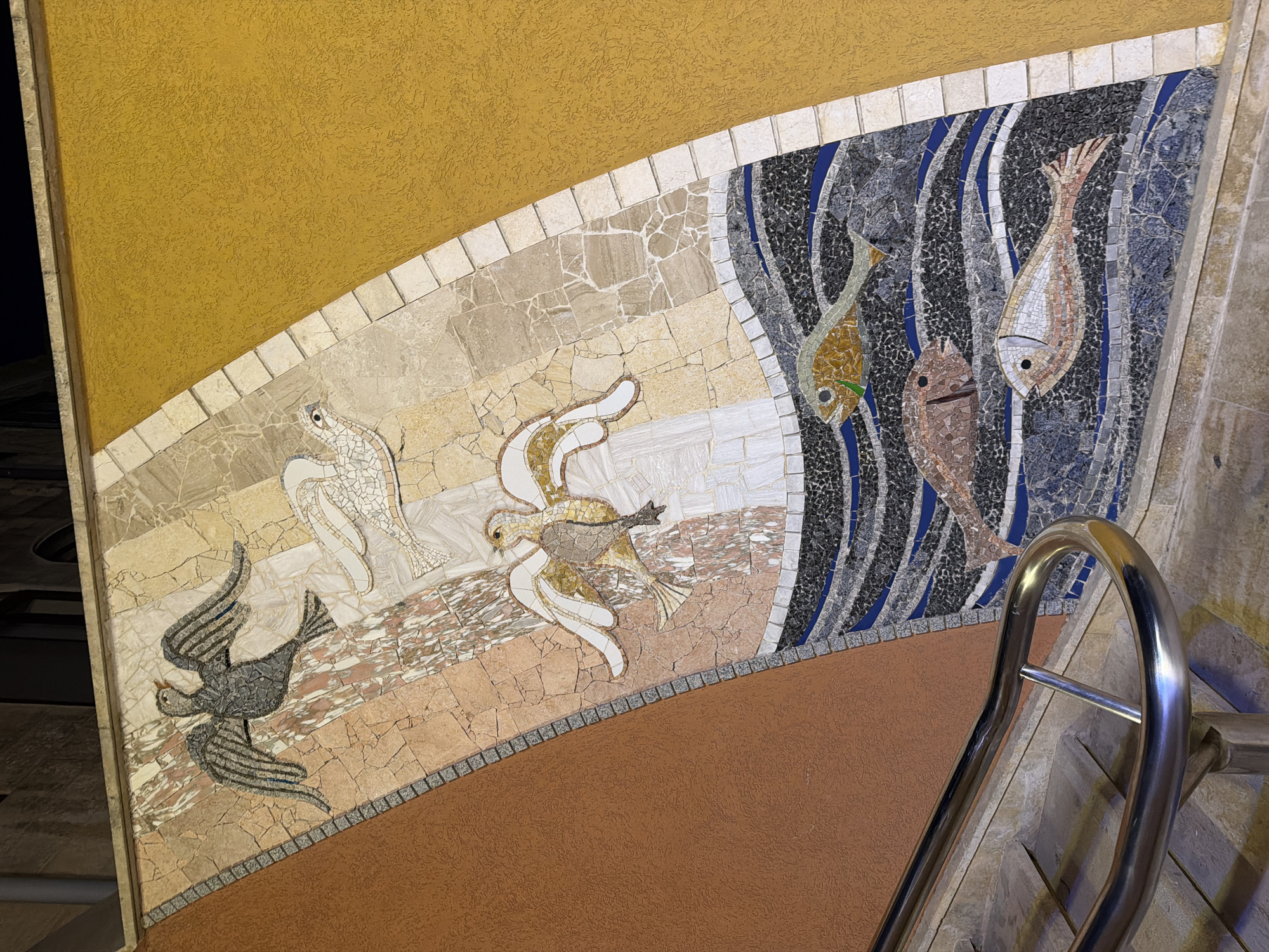 The image shows a mosaic on a wall depicting birds and fish. The mosaic is located on a curved wall next to a staircase with a metal handrail. The mosaic features three birds with outstretched wings and three fish swimming in stylized waves. The birds are depicted in various shades of grey, white, and brown, while the fish are rendered in mosaics of gold, brown, and white. The background of the mosaic is composed of a mix of textured plaster in orange and yellow, and segmented stone tiles. The scene is illuminated by artificial light, creating reflections on the handrail. This mosaic is located in Victoria, Malta, specifically on the facade of the Gozo Museum of Archaeology.