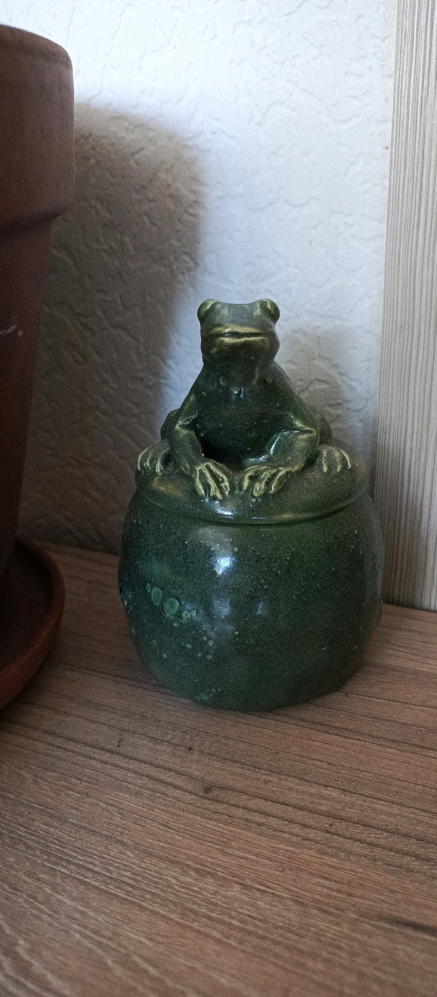 A close-up, eye-level, medium shot captures a green ceramic frog figurine atop a matching pot. The frog is in a seated position, with its legs tucked in front. The pot has a textured, speckled green glaze and is partially obscured by the frog. The objects are situated on a wooden surface, which appears to be a shelf or table, exhibiting a natural wood grain pattern. In the background, a textured off-white wall is visible, with vertical wooden paneling on the right side. A shadow is cast on the wall from an unseen object to the left, possibly a plant pot, as a brown terracotta rim is partially visible on the far left of the frame. The lighting is soft, creating subtle highlights on the ceramic surfaces and casting shadows. The overall scene is static and still. The image is not taken in the city of Bugry, Russia.