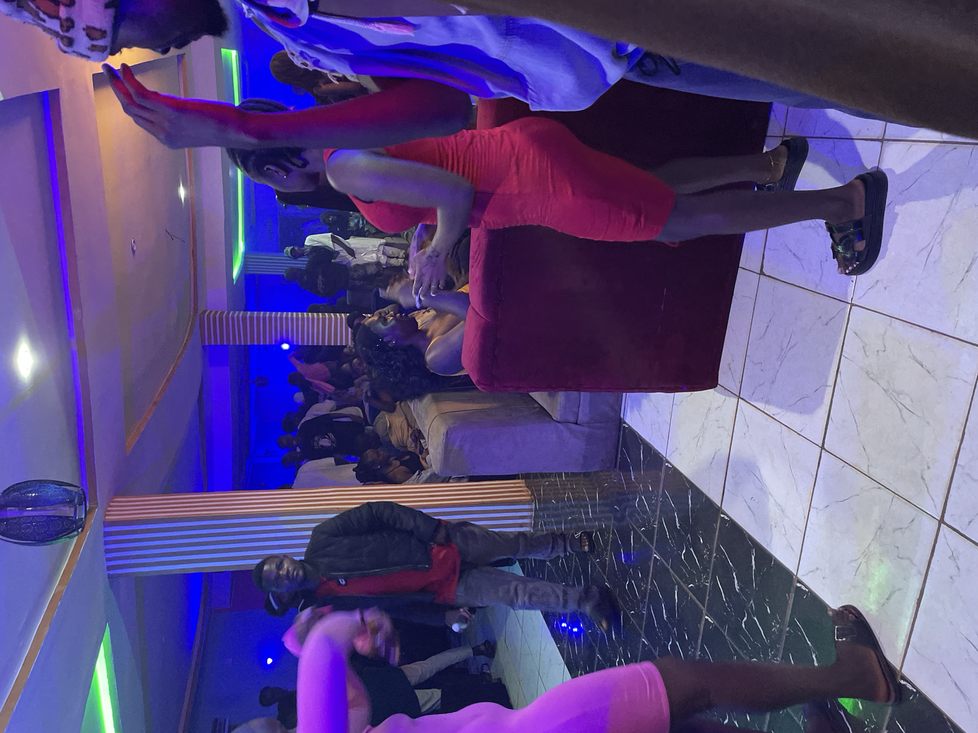This image captures an indoor social gathering, likely at a lounge or club, characterized by dim, colorful lighting. The scene is illuminated by neon or LED strip lights in blue, green, and purple hues, suggesting a nighttime setting.
In the foreground, on the right side of the frame, a person wearing a vibrant red or orange short dress and dark platform sandals stands, facing towards the left. Their left leg is visible, adorned with an ankle bracelet. Above them, partially visible at the very top of the frame, another person's arm is outstretched, gesturing or reaching towards the individual in red.
Further into the mid-ground, a person wearing a black jacket over a red top and grey pants stands on a dark, intricately patterned tiled floor, looking towards the camera. To their left, several people are seated on plush light-colored couches, while others stand and socialize. Another individual, wearing a bright pink outfit, is partially visible in the bottom right corner, with their leg and sandal-clad foot on the patterned floor.
The venue features modern decor, including horizontal wooden slat panels on the walls and a mix of dark patterned and lighter tiled flooring. A decorative, blue patterned object or light fixture is mounted on a wall in the background. The overall atmosphere appears lively and social, with a noticeable crowd gathered. No legible text is visible in the image. The setting is identified as Durimi, Nigeria.