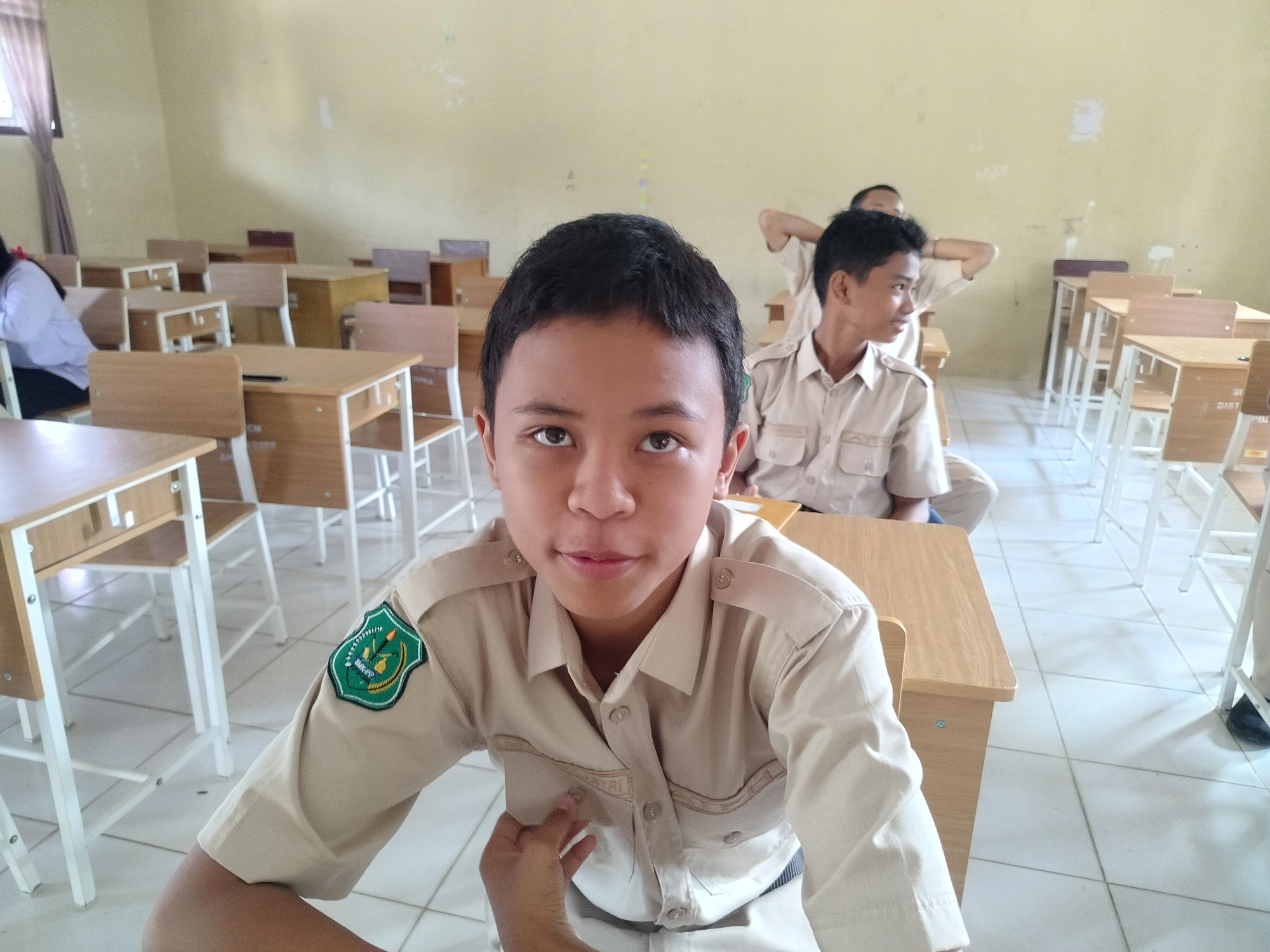 The image depicts a classroom scene, likely in a school in Lawebekung, Indonesia, filled with students in uniform. In the foreground, a young boy with dark hair and brown eyes is looking directly at the camera, wearing a light brown uniform shirt with a green emblem on the left sleeve. His left hand is partially visible, touching his shirt. Behind him, other students are seated at desks. One student in the middle ground is seen from the side, also in a similar uniform, with his hands behind his head. Further back, more students and desks are visible. The classroom has light-colored walls and a tiled floor. Desks are made of wood with white metal frames. A window with a brown curtain is visible on the left side of the frame.