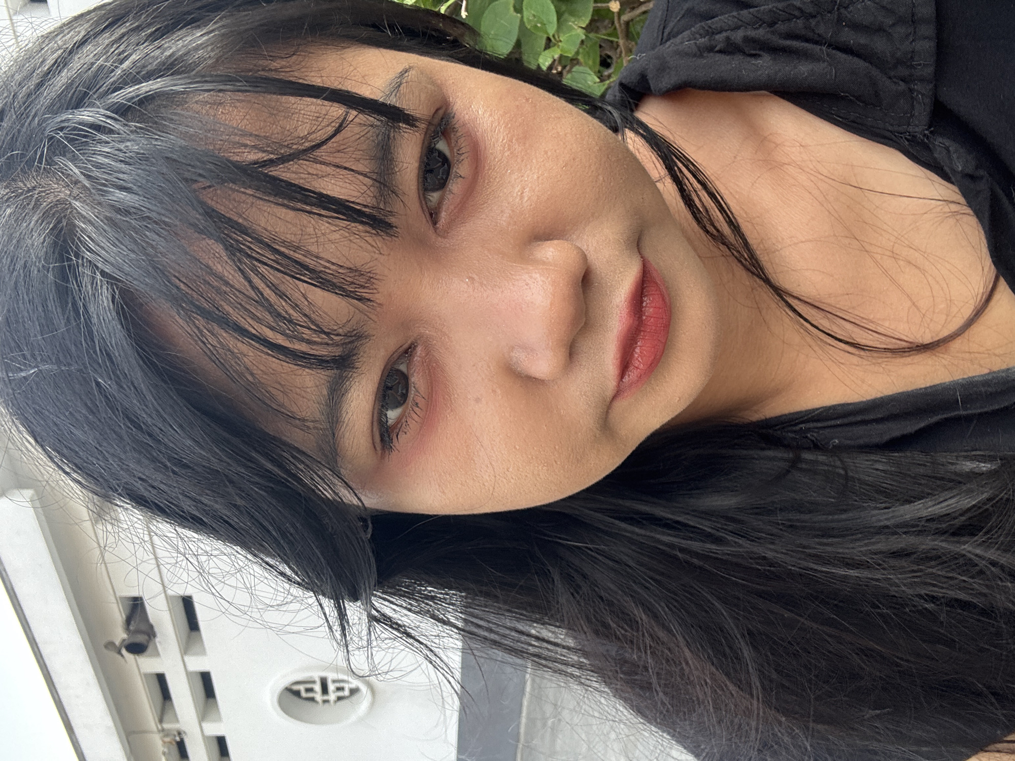 A close-up, front-facing shot captures a young woman with long, dark hair and a black collared shirt. She has visible makeup, including red lipstick and reddish eyeshadow, with some smudging around her eyes. Her skin appears slightly oily, and a few strands of hair are falling across her face. In the background, a white building with a circular decorative element and horizontal window-like openings is visible, suggesting an urban setting. The overall lighting is natural and somewhat overcast.