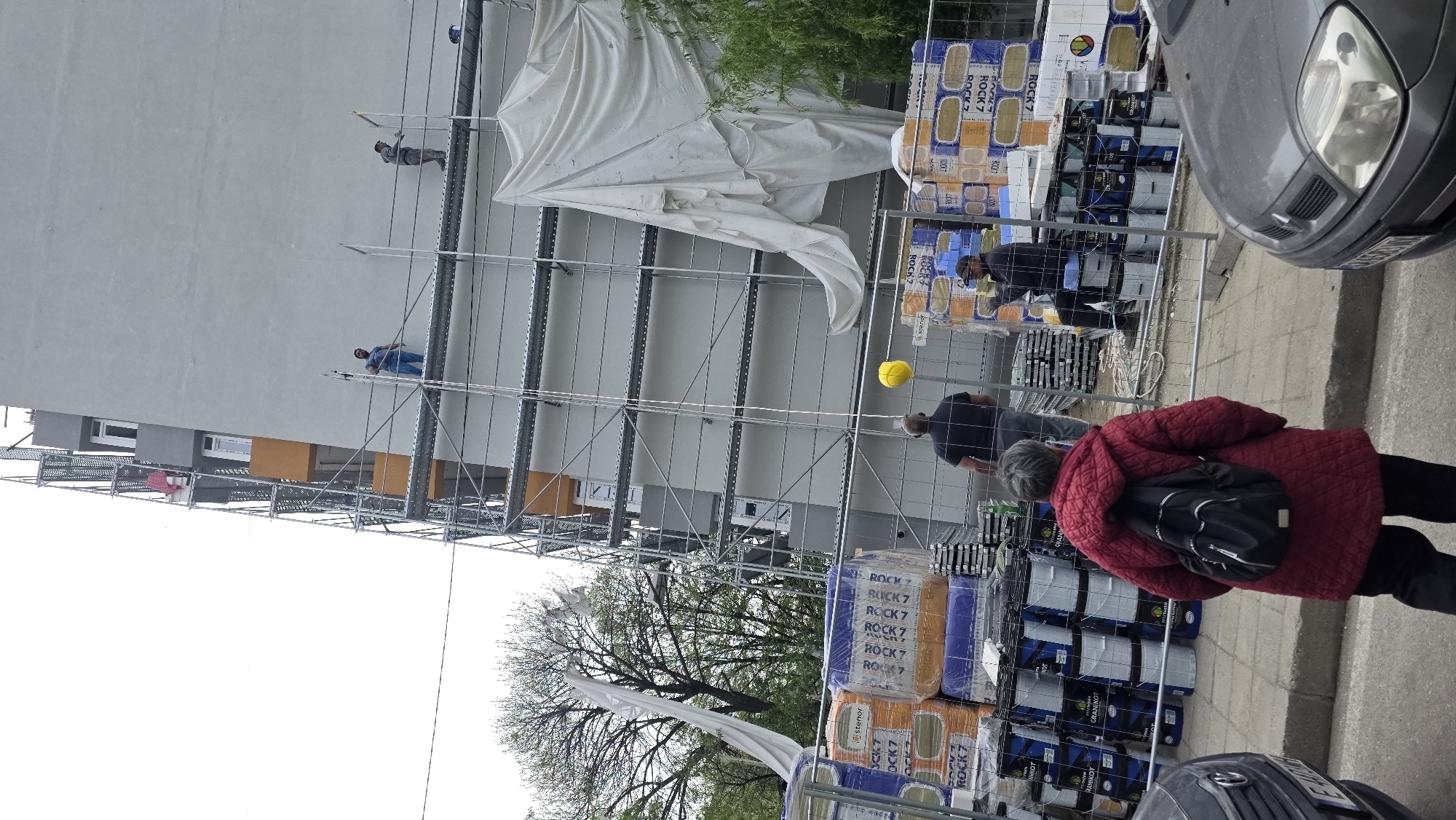The image depicts a construction site scene in an urban area, possibly Blagoevgrad, Bulgaria. A tall building is undergoing renovation or construction, as indicated by the scaffolding erected against its facade. Two workers can be seen on different levels of the scaffolding, one in blue and the other in gray.
In the foreground, stacks of materials labeled "ROCK 7" are visible, along with paint cans or buckets of "GRANIKOT". A woman in a red jacket with a black backpack is walking along the sidewalk in front of the construction site. A yellow hard hat is also visible nearby. A gray car is parked at the right edge of the frame. A portion of the building is covered by a white sheet. Some foliage, including trees, is visible in the background.