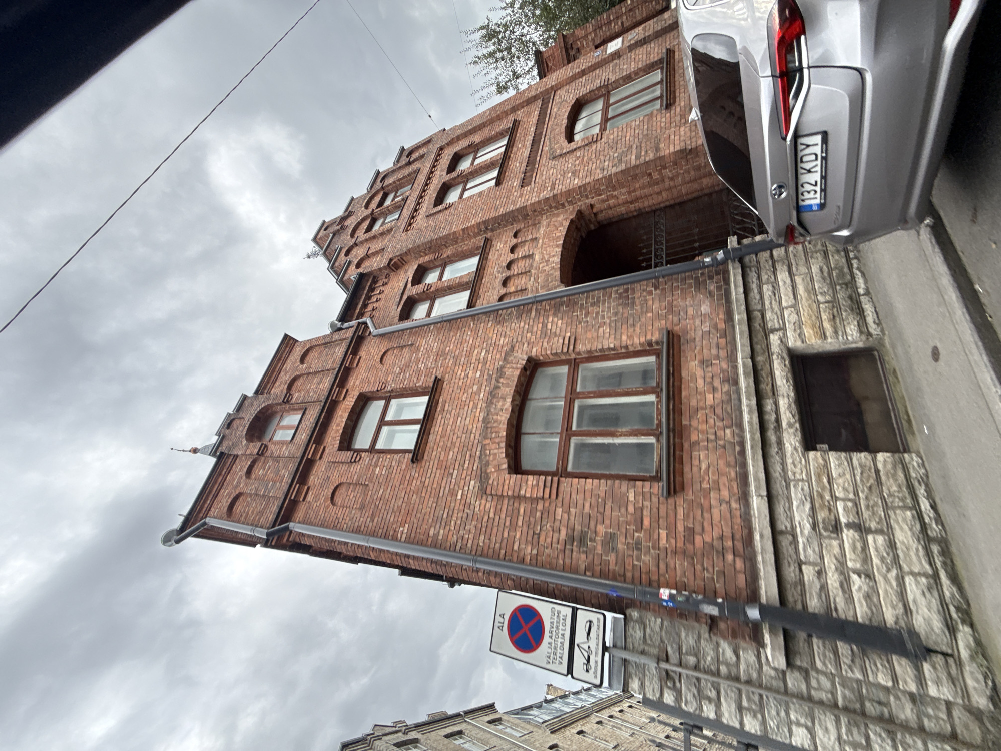 This image captures a street scene in Tallinn, Estonia, focusing on an old brick building with a distinctive stepped gable roof. The building, constructed with aged red bricks, features several arched windows with dark frames. A drainpipe runs vertically along the side of the building. In the foreground, the rear of a silver Toyota sedan is visible, displaying the license plate "132 KDY". A street sign with a red circle and diagonal line over a blue cross is mounted on a pole to the left of the building. The sky is overcast with heavy clouds. The ground appears to be paved, with a curb visible. The overall atmosphere suggests a slightly damp and cool day in the city.