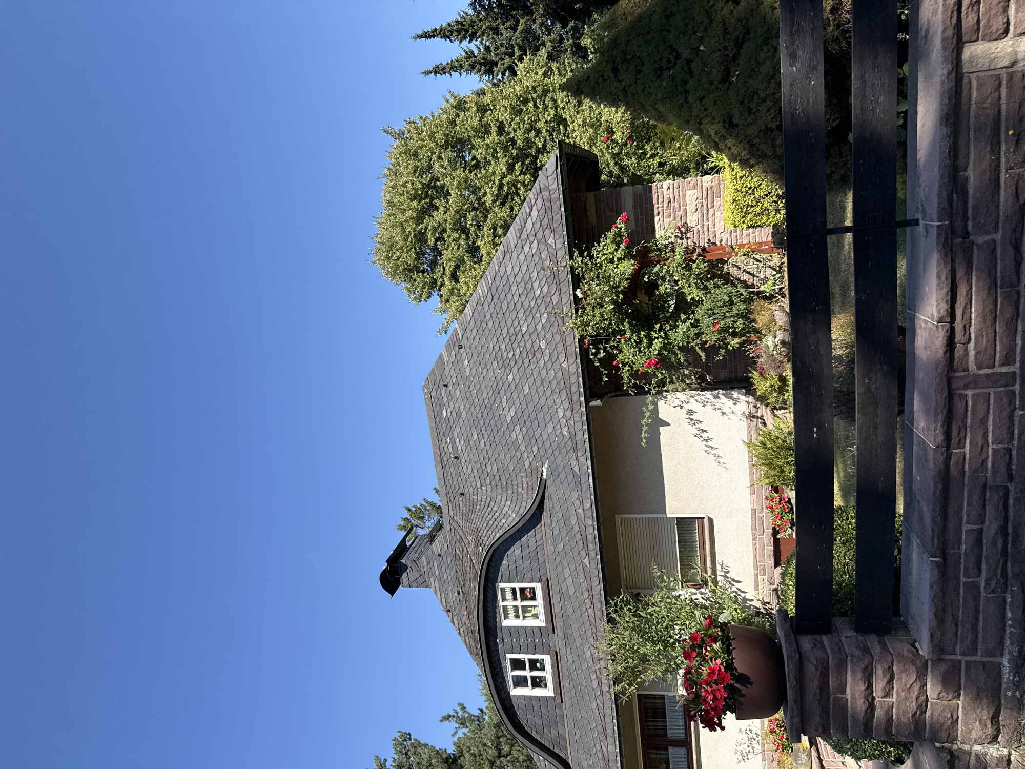 This is a side view of a charming, two-story house with a distinctive mansard roof covered in dark grey slate tiles. The house has cream-colored siding and stone accents. Two white-framed dormer windows are visible on the front-facing slope of the mansard roof. A black chimney with a curved metal cap stands prominently on the roof. The house is surrounded by lush greenery, including mature trees and manicured bushes. A dark wooden fence with stone posts marks the boundary of the property. In the foreground, a large terracotta pot overflows with vibrant red flowers. The sky above is a clear, bright blue. This scene appears to be in a residential area of Bad Sachsa, Germany, characterized by its well-maintained properties and natural beauty. There are no people or animals present in the image.