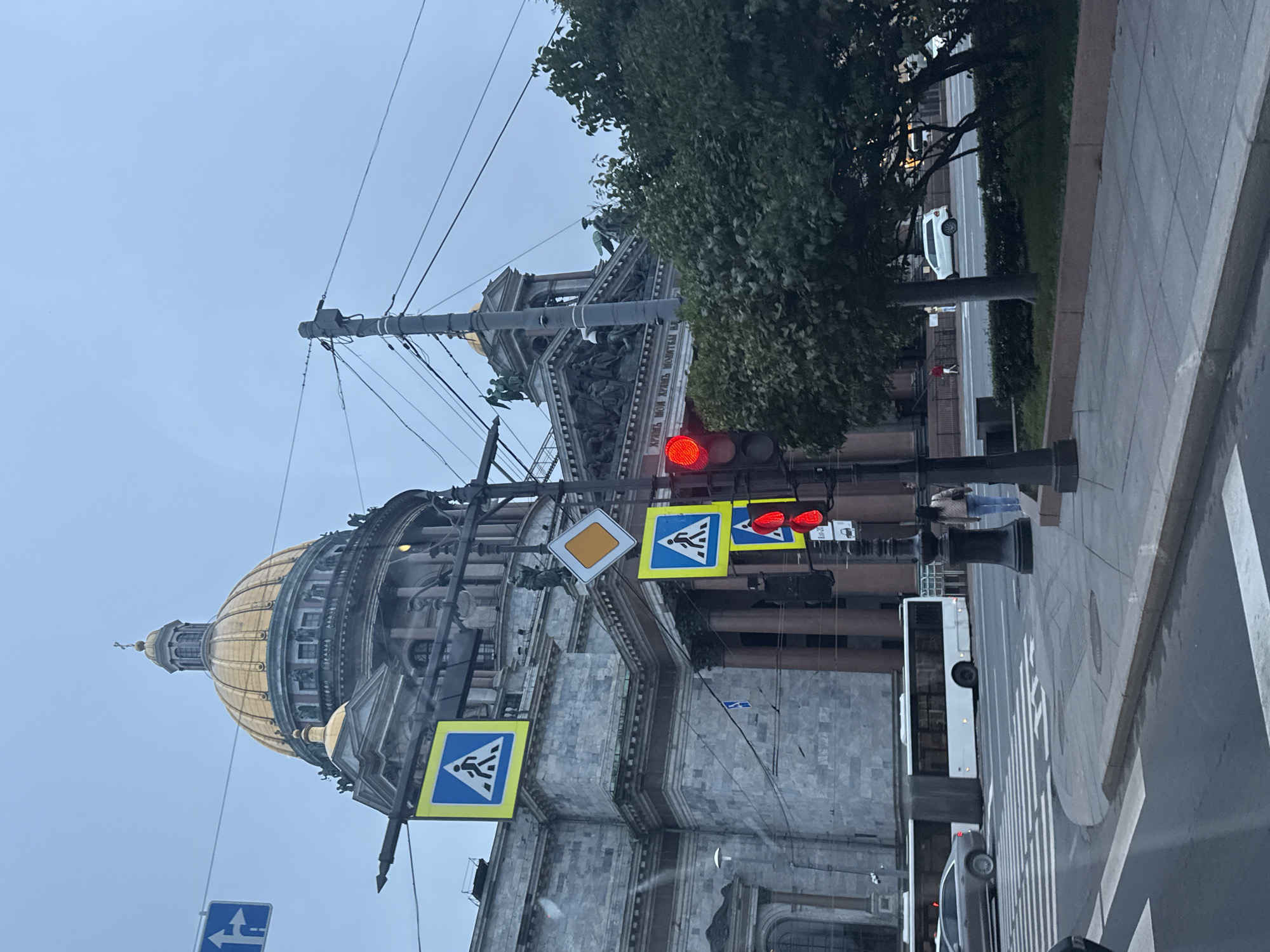 The image depicts a view of Saint Isaac's Cathedral in Saint Petersburg, Russia, on a cloudy day. In the foreground, a street with a pedestrian crossing is visible, along with a traffic light showing red. Several traffic signs, including pedestrian crossing signs and a directional arrow, are also present. A white bus and a silver car are seen on the road. A woman in a light-colored jacket and blue jeans is walking across the street. To the right, a dense cluster of trees partially obscures the view of the cathedral's facade. In the background, the majestic dome and exterior of Saint Isaac's Cathedral are prominent. The overall atmosphere is overcast, suggesting a cool or temperate climate.