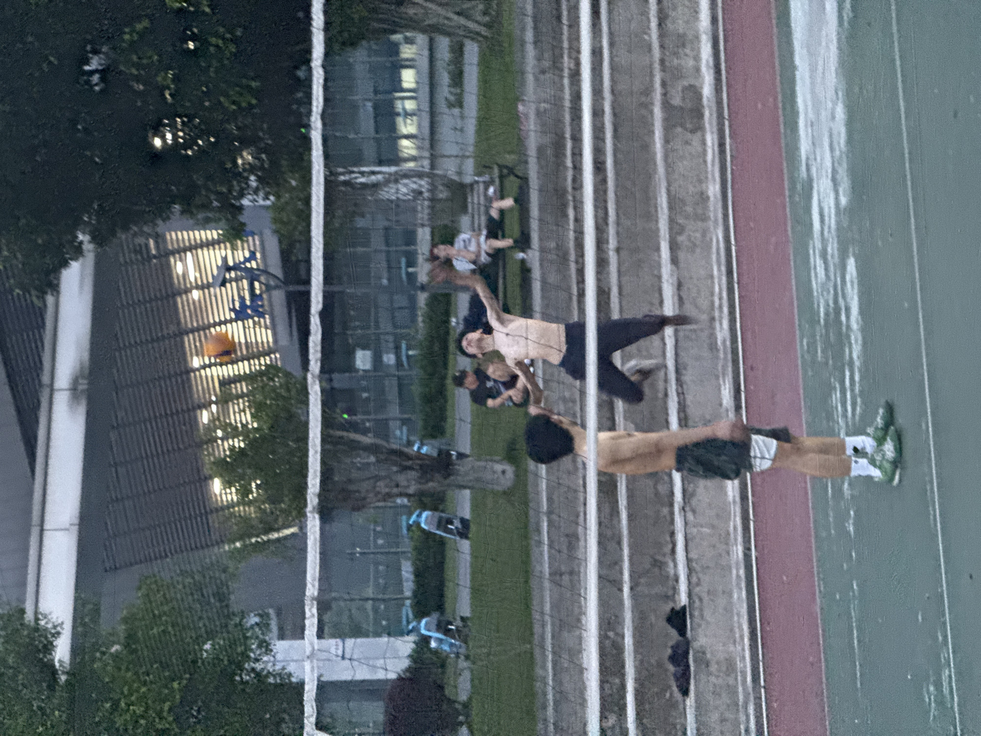 This image depicts an outdoor sports court scene in Jiangjin, China, during dusk or early evening. The primary focus is on a group of individuals engaged in recreational sports. In the mid-ground, a shirtless man with dark shorts is captured in a dynamic pose, leaping or reaching over a white sports net, likely playing volleyball. Closer to the camera, another shirtless man in dark shorts and light sneakers stands near the net, with his back partially to the viewer.
Further in the background, several other people are present, observing or participating. One individual in a light-colored top is seated or resting on the grassy area. To the left of the court, a basketball hoop with an orange net is visible, and a basketball lies nearby, indicating the presence of a multi-sport facility. The court itself features a distinct reddish-brown strip adjacent to a greenish-gray surface. Behind the sporting area, modern buildings with vertically patterned facades and illuminated windows rise, their lights reflecting in the dimming light. Green trees are interspersed throughout the background. The scene conveys a relaxed, casual atmosphere of outdoor leisure and exercise. There is no visible text in the image.