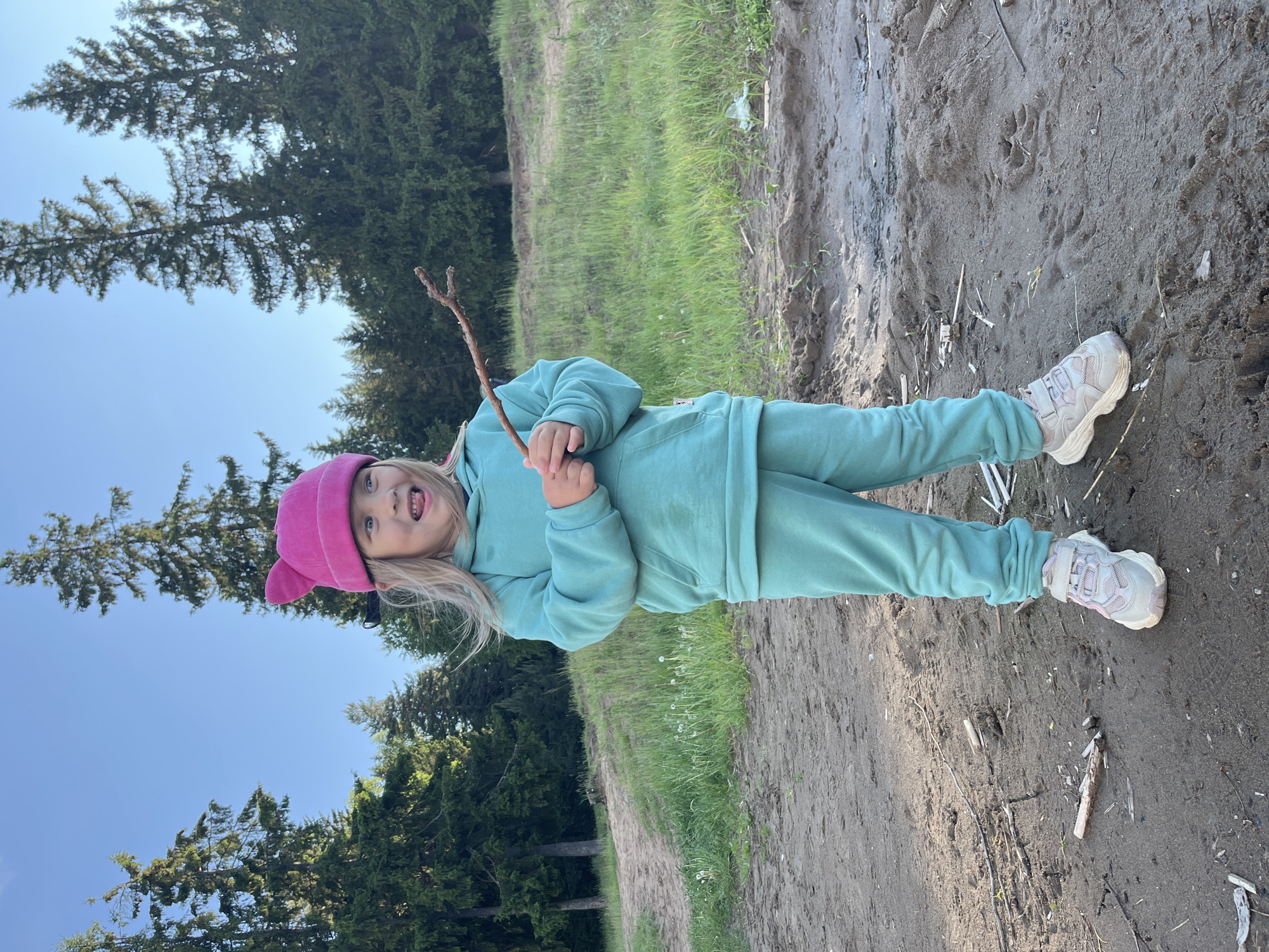 A young child is depicted outdoors, standing on a sandy or dirt path. The child, who appears to be a girl with blonde hair, wears a teal-colored sweatsuit and a bright pink hat with ear-like features. She holds a small, forked stick in both hands and has an open-mouthed, seemingly playful or excited expression directed upwards and slightly to the left.
The setting is a natural outdoor environment, likely a forest edge or a recreational area in Chaikovsky, Russia. To the child's left, there is a grassy slope, and behind her, dense green conifer trees rise towards a clear, bright blue sky, suggesting sunny weather during daytime, possibly late morning or early afternoon. The ground is composed of sand or dirt, with scattered small twigs and debris. Near the child's right foot, a distinct animal paw print is visible in the sandy surface. In the background, on the dirt path, a discarded white plastic bag can be faintly seen. The overall scene conveys a moment of childhood play and exploration in a natural, serene environment. No visible text is present in the image.