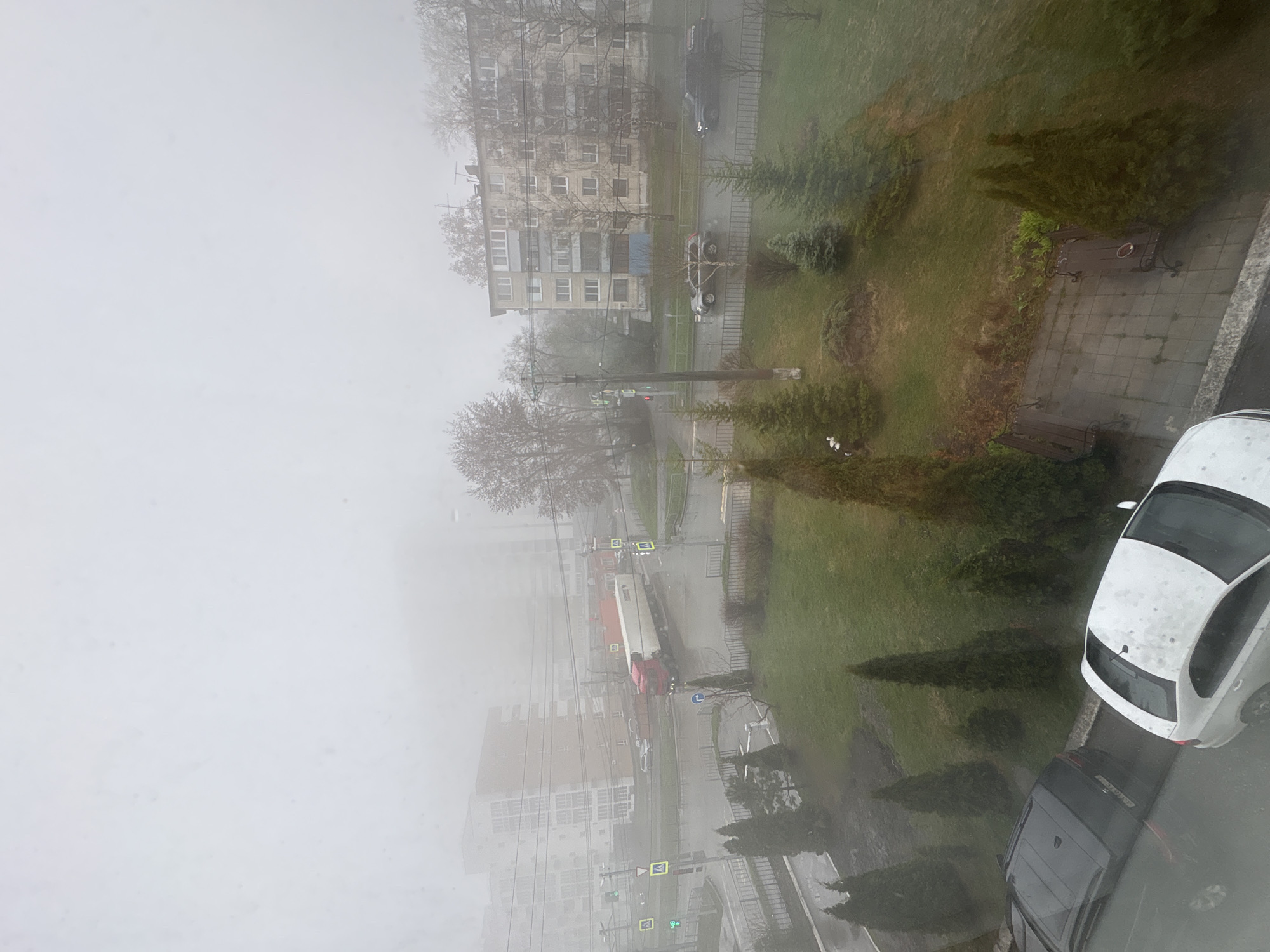 This image captures an urban street scene shrouded in dense fog, viewed from a high vantage point, likely an apartment window. The pervasive fog dramatically reduces visibility, giving the entire landscape a muted, low-contrast, and hazy appearance.
In the foreground on the right, a white hatchback car, possibly a Volkswagen Golf or Polo, is prominently parked, its surface speckled with water droplets. Behind it, a dark-colored vehicle is partially visible. Below these cars, a paved walkway featuring two dark park benches runs alongside a grassy area dotted with various evergreen shrubs and deciduous trees, many of which are bare, suggesting late winter or early spring.
Towards the center and left of the frame, a road meanders through the scene. A red and white semi-trailer truck is visible on this road, facing away from the viewer. Traffic lights, utility poles with overhead wires, and several square, yellow-bordered pedestrian crossing signs are discernible along the roadside, though details are softened by the mist.
In the middle ground, an older, multi-story apartment building with numerous windows and balconies stands partially obscured by the fog and the bare branches of trees. Further down the street, newer, taller buildings are faintly visible, also enveloped in the thick fog. The sky is entirely white and indistinct due to the heavy mist, preventing a clear determination of the time of day, though it is clearly daylight. The wet surfaces on cars and pavement indicate recent rain or extremely high humidity. No specific activity or event is taking place, and no people are distinctly visible. The scene conveys a quiet, somewhat somber atmosphere typical of a heavily foggy day in a residential area of Ulyanovsk, Russia.