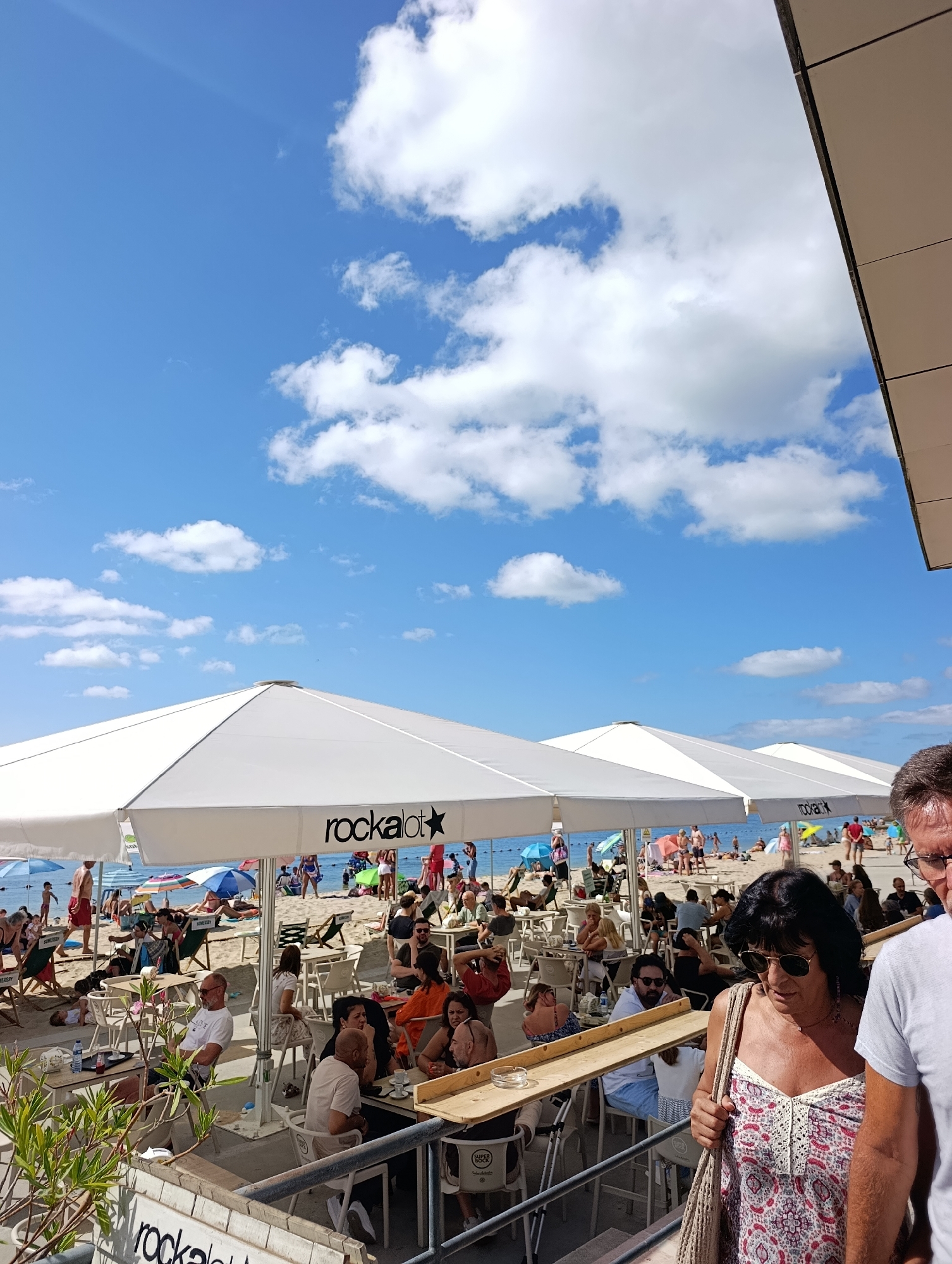The image depicts a crowded outdoor seating area of a beachside establishment in Setúbal, Portugal, likely during daytime under a clear blue sky with scattered white clouds. Several white, hexagonal umbrellas providing shade for tables and chairs are prominent. The umbrellas feature the "rockalot" logo. In the background, a beach with numerous sunbathers, umbrellas, and the ocean is visible. People are seated at tables, some are walking, and others are lounging on the beach. The foreground shows more seating areas with people engaged in various activities, such as eating, drinking, and conversing. The overall atmosphere suggests a lively and popular recreational spot.