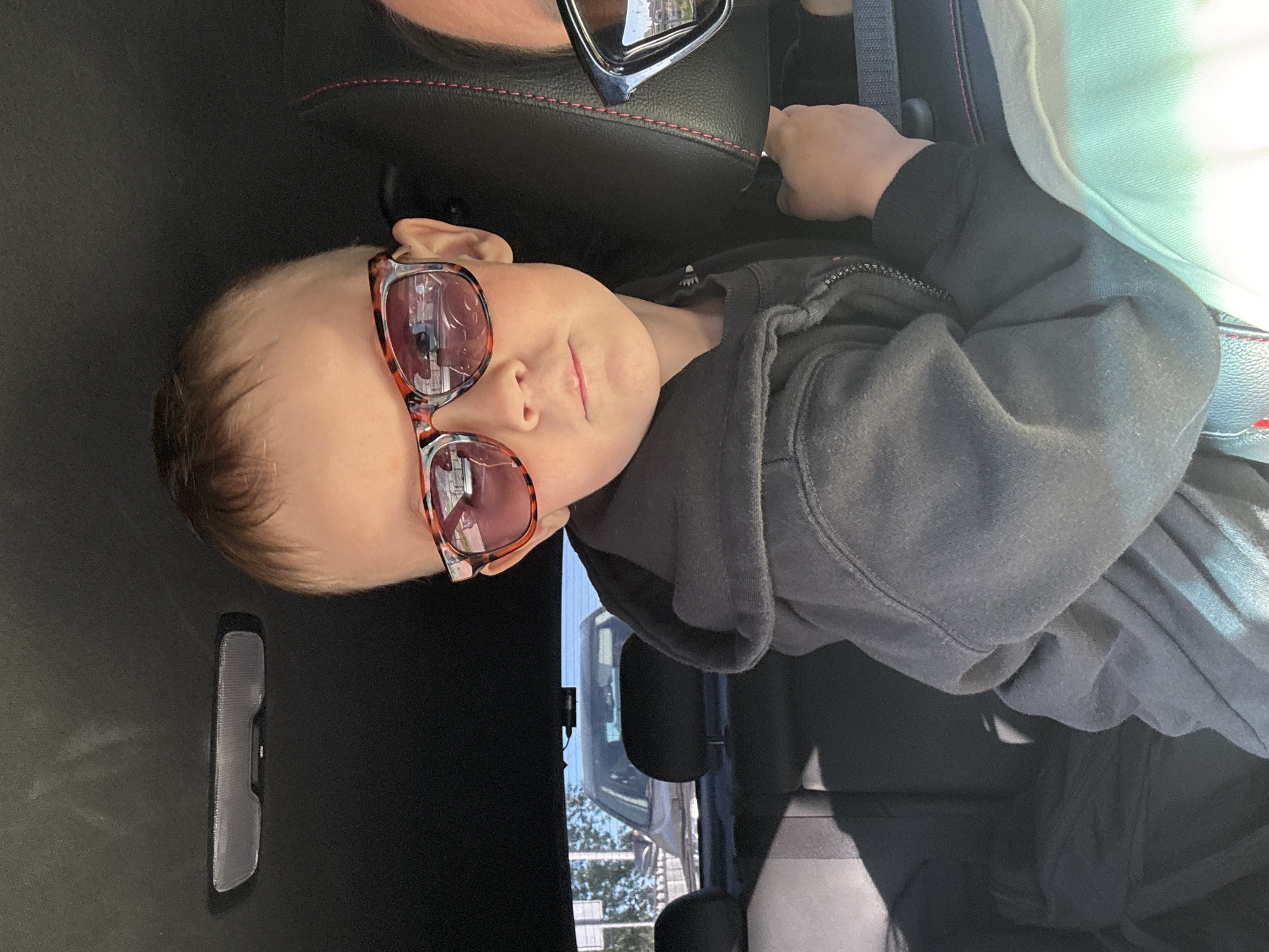 A young child wearing sunglasses sits in the backseat of a car. The interior of the vehicle is predominantly black, with a gray hooded sweatshirt worn by the child. The child is facing forward with a slight tilt to their head. Outside, through the car window, the blurred image of another vehicle and some trees are visible. The location, Chaikovsky, Russia, cannot be determined from the given information.