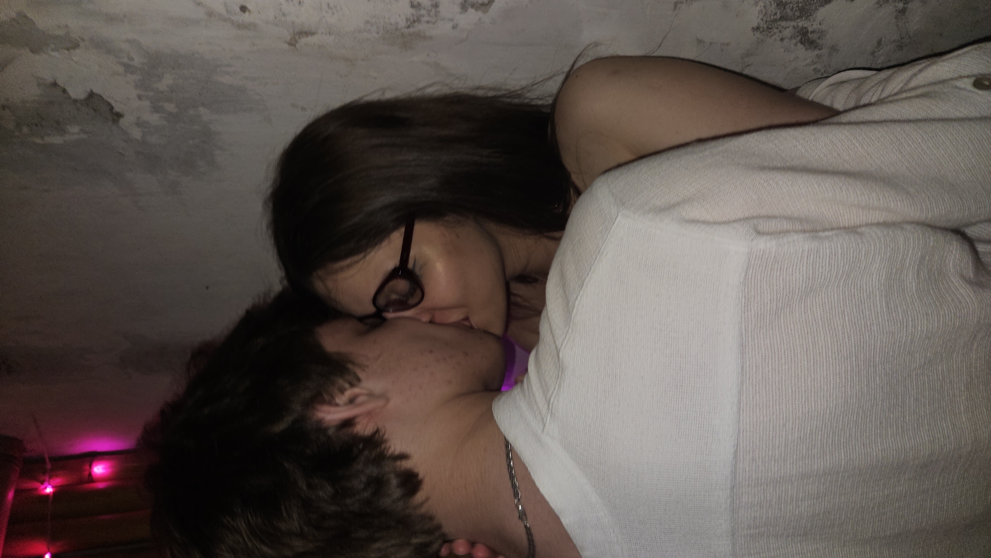 Two individuals are kissing. A fair-skinned woman with long brown hair and wearing glasses is on the upper left, leaning over a man with short brown hair. The man has a chain necklace. They are indoors, with a textured wall visible above. There are pink string lights in the lower left corner. The woman is wearing a white shirt. The location is unknown and cannot be determined from the image.