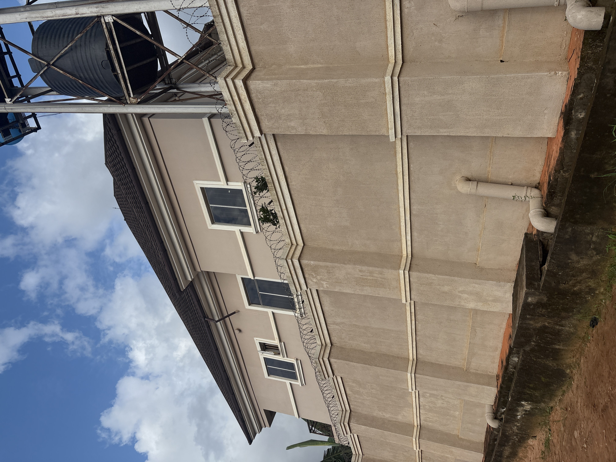 The image shows a side view of a two-story building with a beige-colored facade, situated in Obosi, Nigeria. The building has a dark brown tiled roof and several windows, some of which are open. The property is enclosed by a tall, beige-colored wall topped with barbed wire fencing. In the background, a large black water tank stands on a metal structure. The sky above is blue with scattered white clouds. The foreground shows a dirt path and a concrete drainage channel. The overall scene suggests a residential or commercial property with security measures in place.