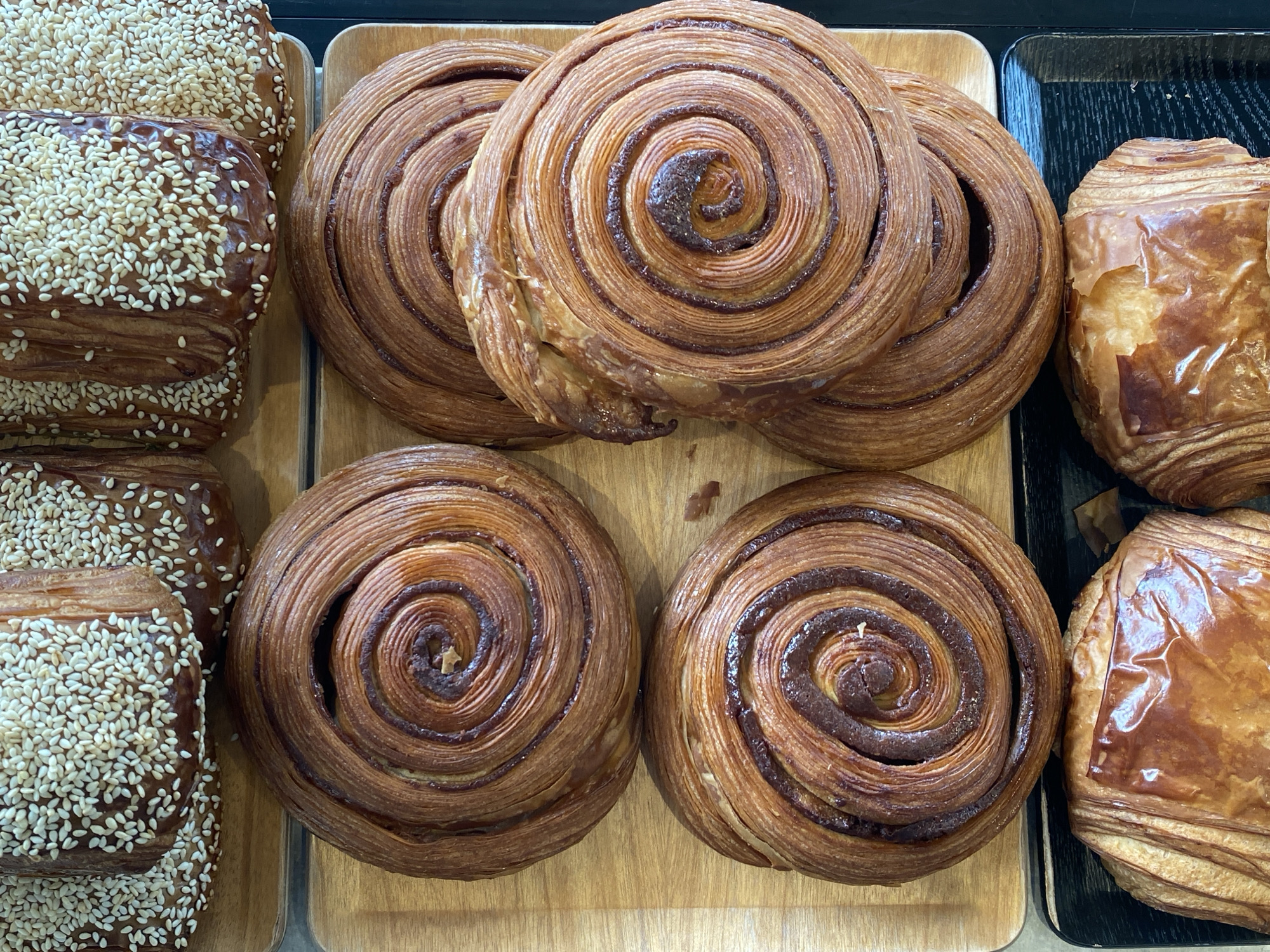 The image shows a selection of pastries on display, likely in a bakery or café. The pastries are arranged on wooden and black trays. On the left are several sesame-seed-covered pastries. In the center are four cinnamon rolls with a swirled pattern. On the right are two croissant-like pastries with a golden brown glazed surface. The pastries are close together, indicating that they are ready for purchase. Given the location, this is likely from Tel-Aviv, Israel.
