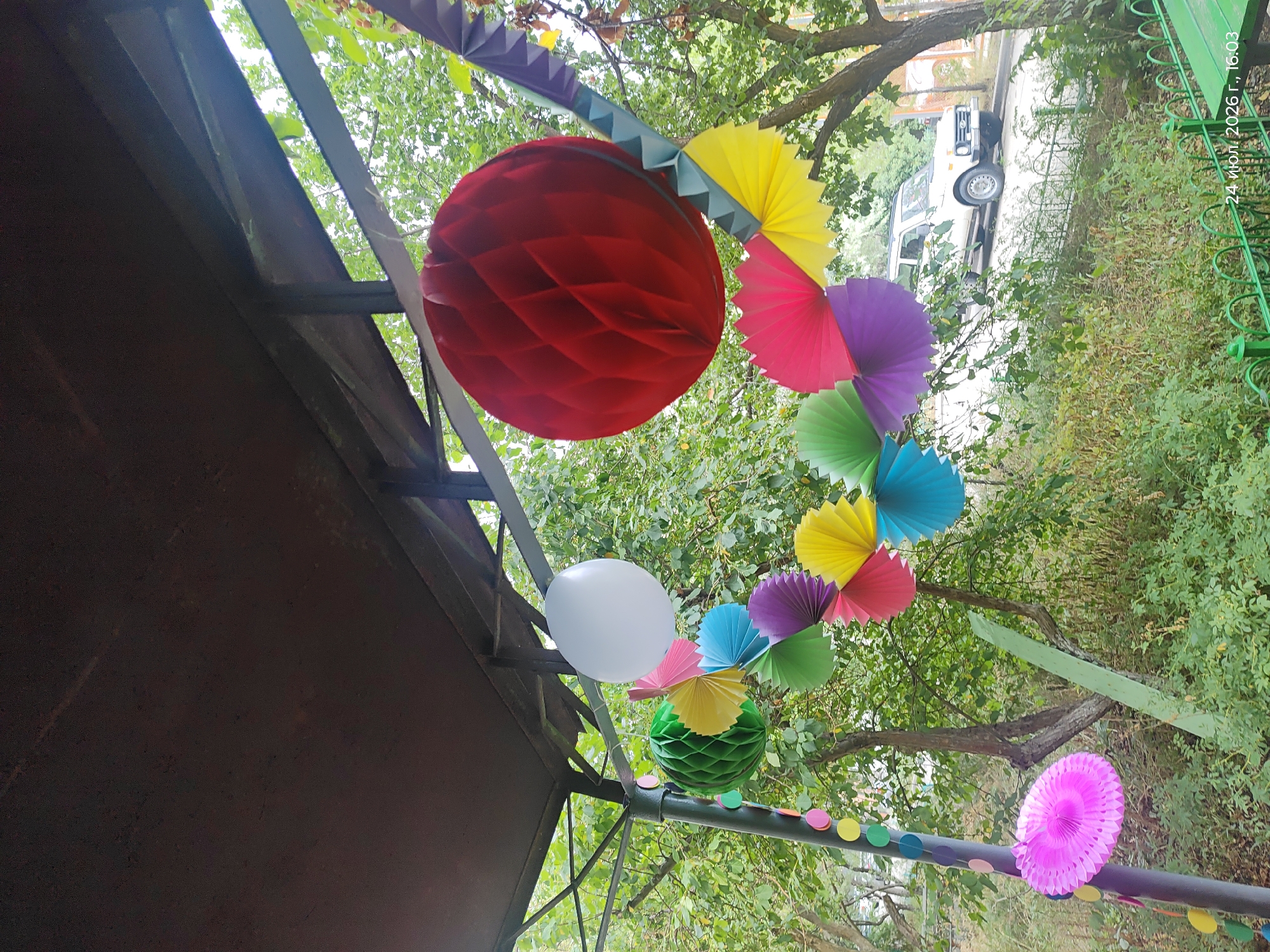 The image depicts an outdoor scene under a dark, weathered roof structure, likely a veranda or overhang, possibly in a park or backyard. A string of colorful paper decorations, including round honeycomb balls and fan-shaped paper ornaments in various hues such as pink, yellow, blue, purple, and green, are suspended from the structure. A large red honeycomb ball hangs prominently in the foreground, to the right of a white balloon. A green honeycomb ball is visible to the left. In the background, lush green trees and bushes are present, indicating a natural environment. A white SUV is parked on a road that curves uphill to the right of the frame, partially obscured by foliage. The ground appears to be covered in grass and vegetation. A green metal fence runs along the bottom of the image. The timestamp at the bottom right corner reads "24 июля, 2026 г., 16:03" which translates to "July 24, 2026, 16:03". The mention of Astana, Kazakhstan, is absent from the visual information.