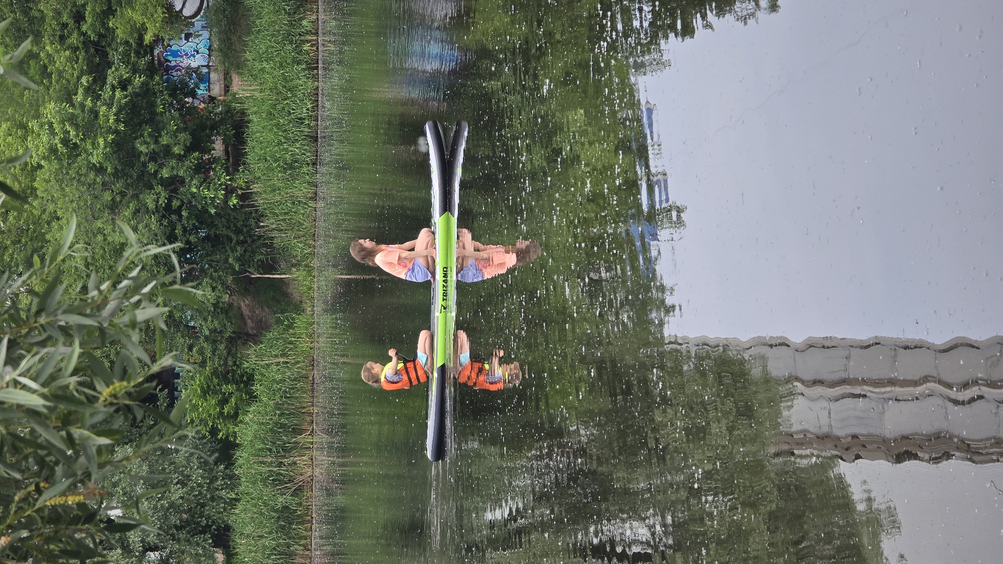 Two girls are paddling on a green and black TRIZANO inflatable stand-up paddleboard on a dark, reflective body of water. The younger girl, on the left, wears an orange life vest over a light top and blue shorts, holding a paddle and looking to her right. The older girl, on the right, wears a pink t-shirt and light blue shorts, seated with her knees bent and also looking to her right. The water's surface shows ripples and numerous small white specks, with clear reflections of the girls, the paddleboard, and the surrounding environment.
In the background, a lush green bank features tall reeds and dense trees. Towards the right side of the bank, a concrete retaining wall is visible, heavily covered with multi-colored graffiti. Overhanging green leaves from a tree are out of focus in the top left corner of the image. The lower left of the water reflects a distorted, wavy image of a tall, light-colored building or structure.
The scene depicts recreational activity on the Rusanivka Channel in Kyiv, Ukraine, an artificial waterway known for its green banks and residential backdrop, where concrete embankments often feature street art.