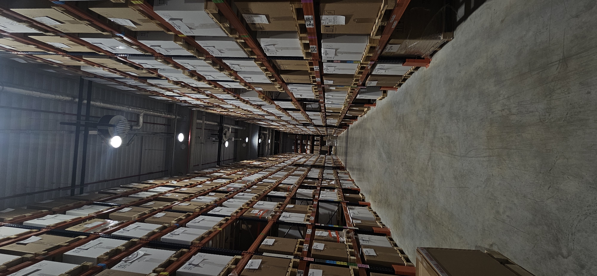The image shows an interior shot of a large warehouse. The floor is concrete. High shelving units, stacked with cardboard boxes, extend on both sides to the far end. Lighting fixtures are visible on the ceiling. The location and city cannot be discerned from the image.