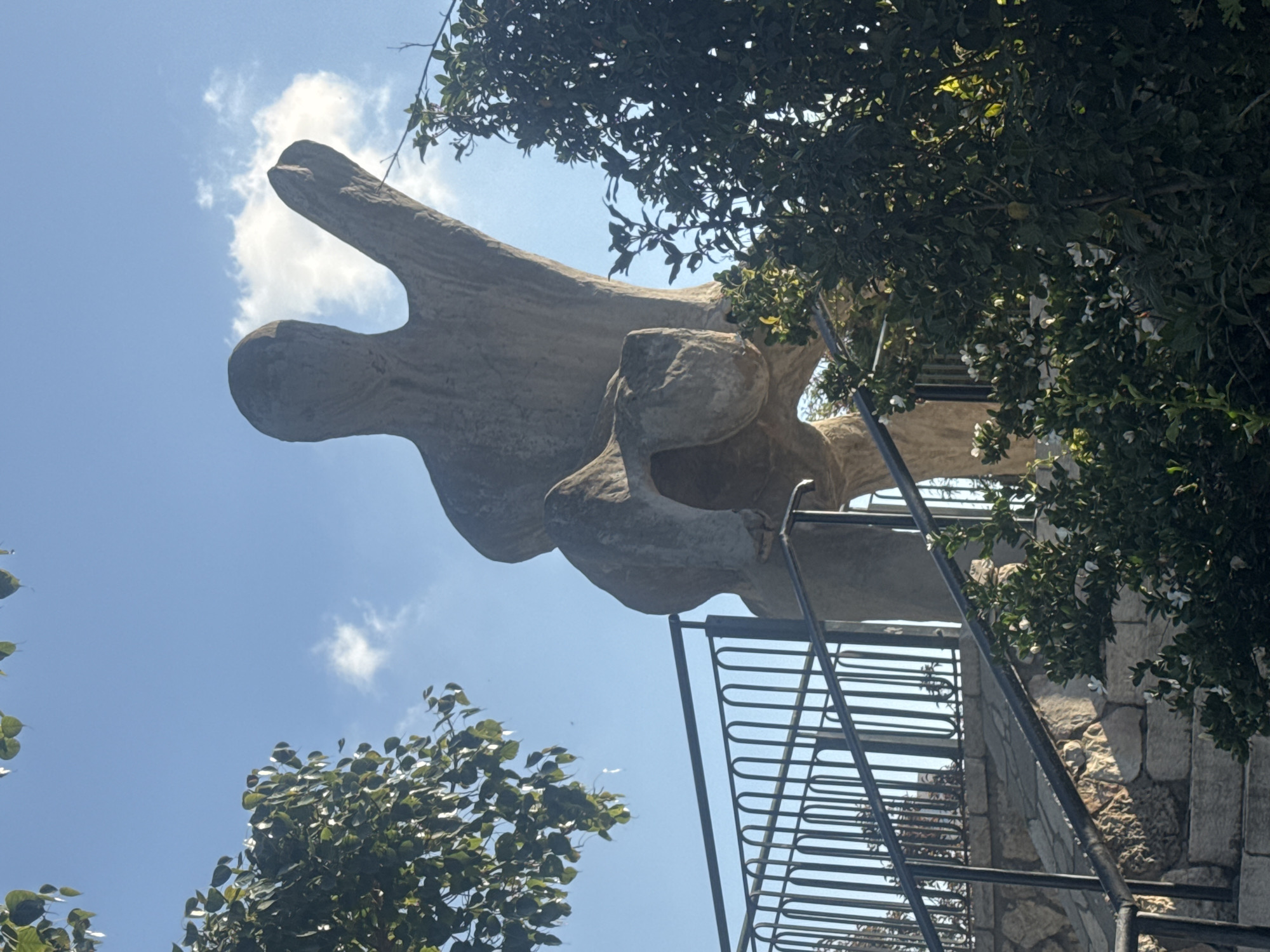 The image shows a sculpture titled "Man in the Void" by Israeli artist Ilan Averbuch. The sculpture, resembling a stylized human figure with an elongated head, is made of light-colored stone. The figure appears to be suspended against a partly cloudy blue sky, viewed from a low angle. The sculpture is located outdoors, and visible elements include parts of a stone structure, a metal ladder, and branches of trees with green leaves and white flowers partially framing the scene. The sculpture is in Ramat Gan, Israel.