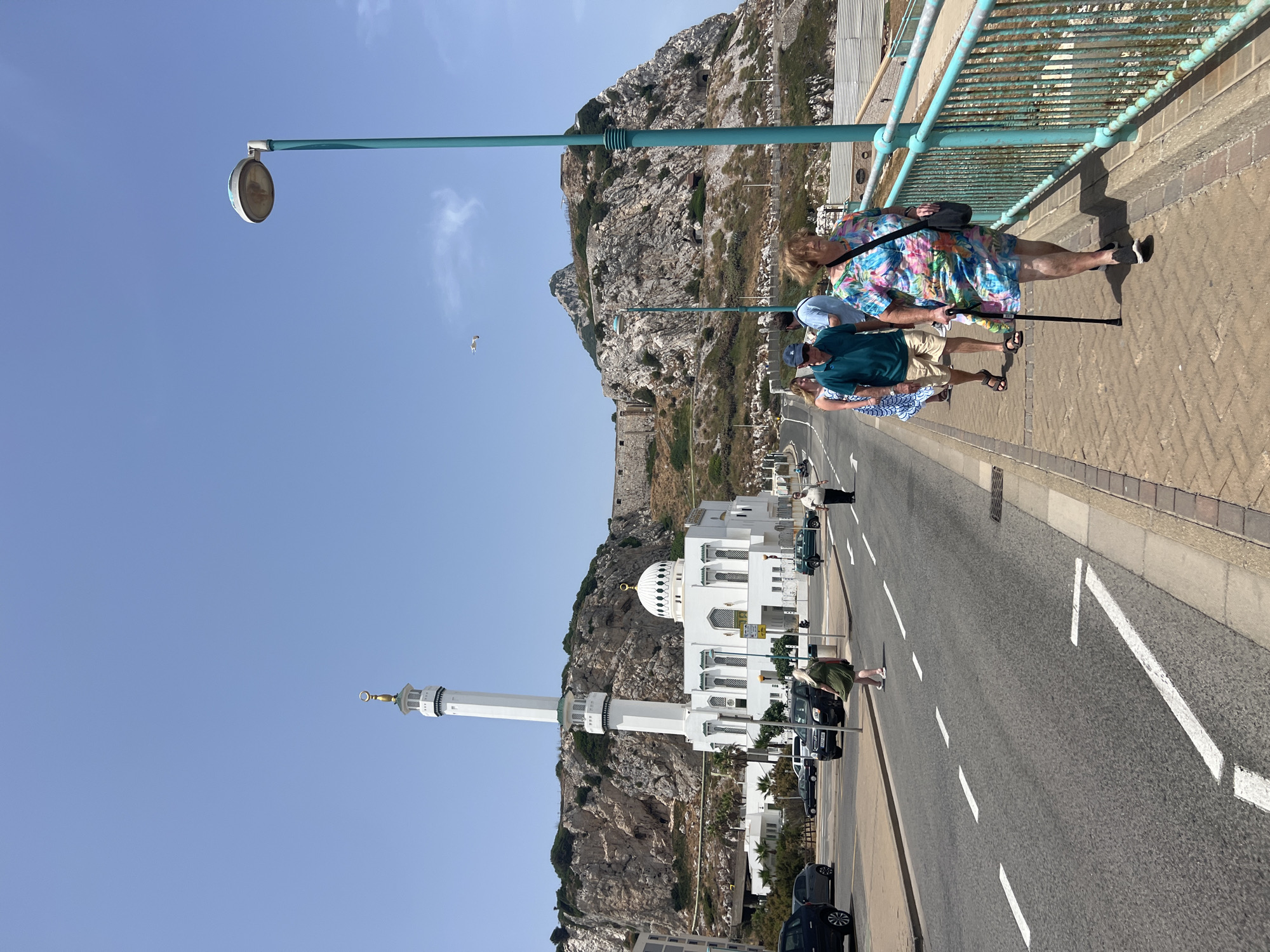 The image shows a street scene in Gibraltar. In the background, the King Fahd bin Abdulaziz Al Saud Mosque and the Rock of Gibraltar are visible. A group of people are walking on a sidewalk next to a road. One woman in the foreground is using a cane and wearing a colorful dress. Cars are parked and driving on the road. The sky is clear and blue.