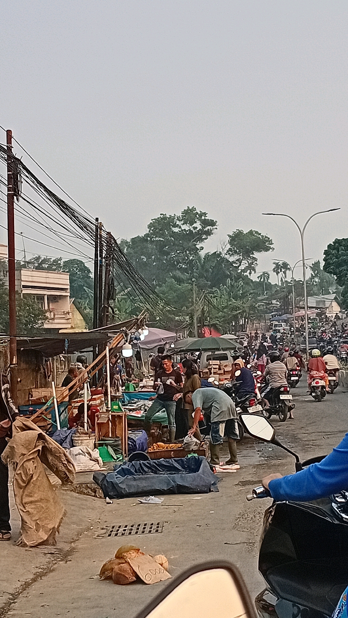 The image depicts a bustling street market scene in Jambi, Indonesia, likely on a cloudy day. The foreground shows a dirt and asphalt road with several parked motorcycles and a person on a motorcycle wearing a blue jacket. Various market stalls are set up along the side of the road, displaying goods that are partially obscured by tarps and awnings. Several individuals are engaged in transactions or tending to their stalls. One man in a gray t-shirt and jeans is seen interacting with a vendor. Another person, wearing a blue shirt and white sneakers, is bent over a stall. Further down the street, a dense crowd of people and motorcycles suggests ongoing commercial activity. The background features a mix of residential buildings, lush green trees, and utility poles with a tangled web of electrical wires. The overall atmosphere is one of lively commerce and everyday life in a Southeast Asian urban setting.