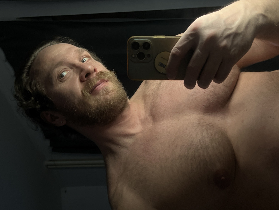 A selfie captures a shirtless man with a full beard and mustache, holding a gold-colored smartphone. The man gazes upward, his curly brown hair visible. The focus is on his chest and upper body, revealing body hair. The backdrop appears dark.