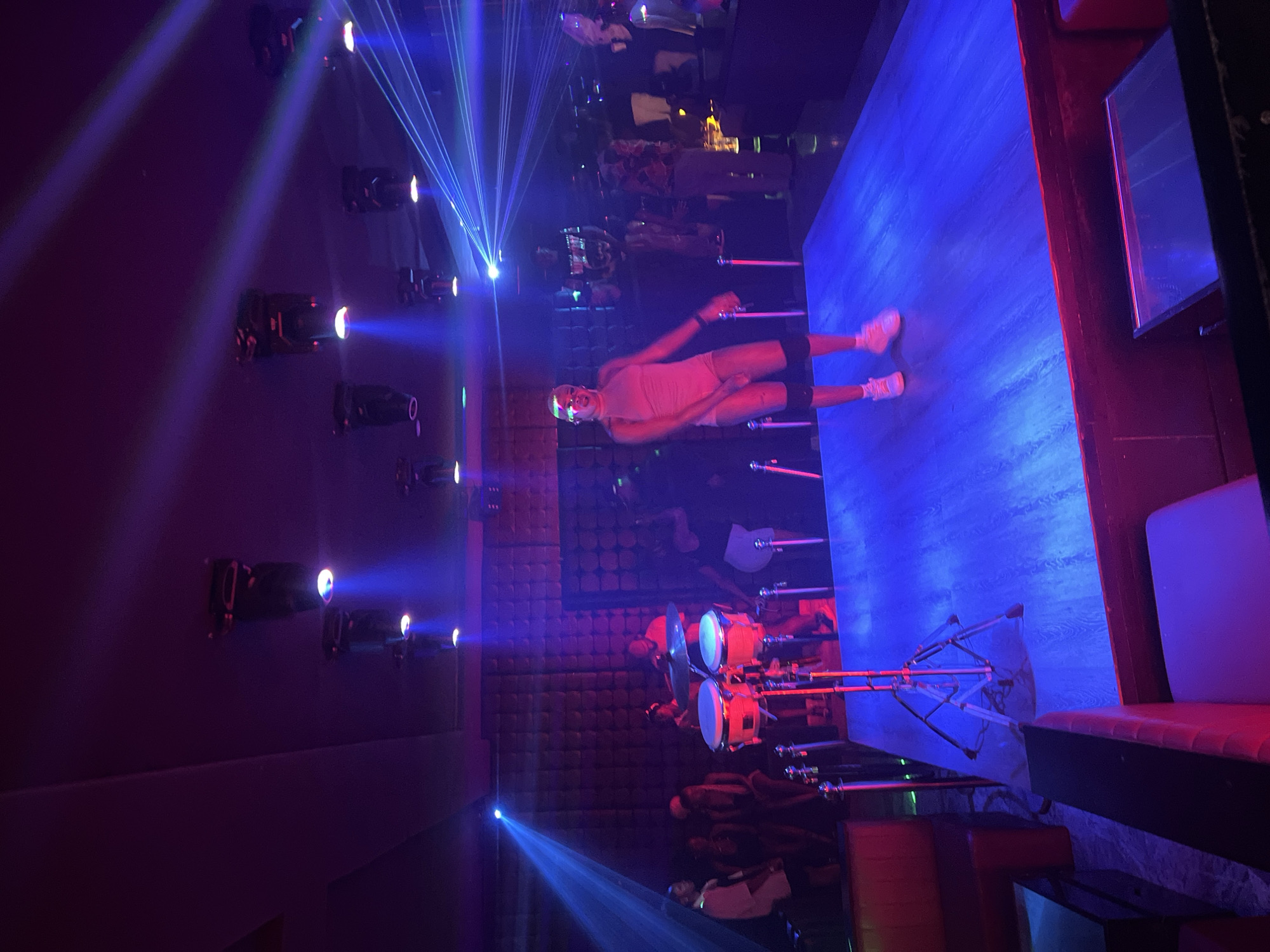 The image depicts a dimly lit nightclub in the city of Howayi, Nigeria, illuminated by vibrant blue and purple stage lights. A person in a pink outfit is dancing on a raised, blue-lit stage. To the left of the stage, a drum set is set up, with a drummer playing it. Several other individuals are visible in the background, some appearing to be patrons of the club, while others might be performers or staff. Laser lights are projected across the ceiling and stage. Red seating is arranged in the foreground and background, suggesting a seating area for guests. The overall atmosphere is that of a lively entertainment venue.