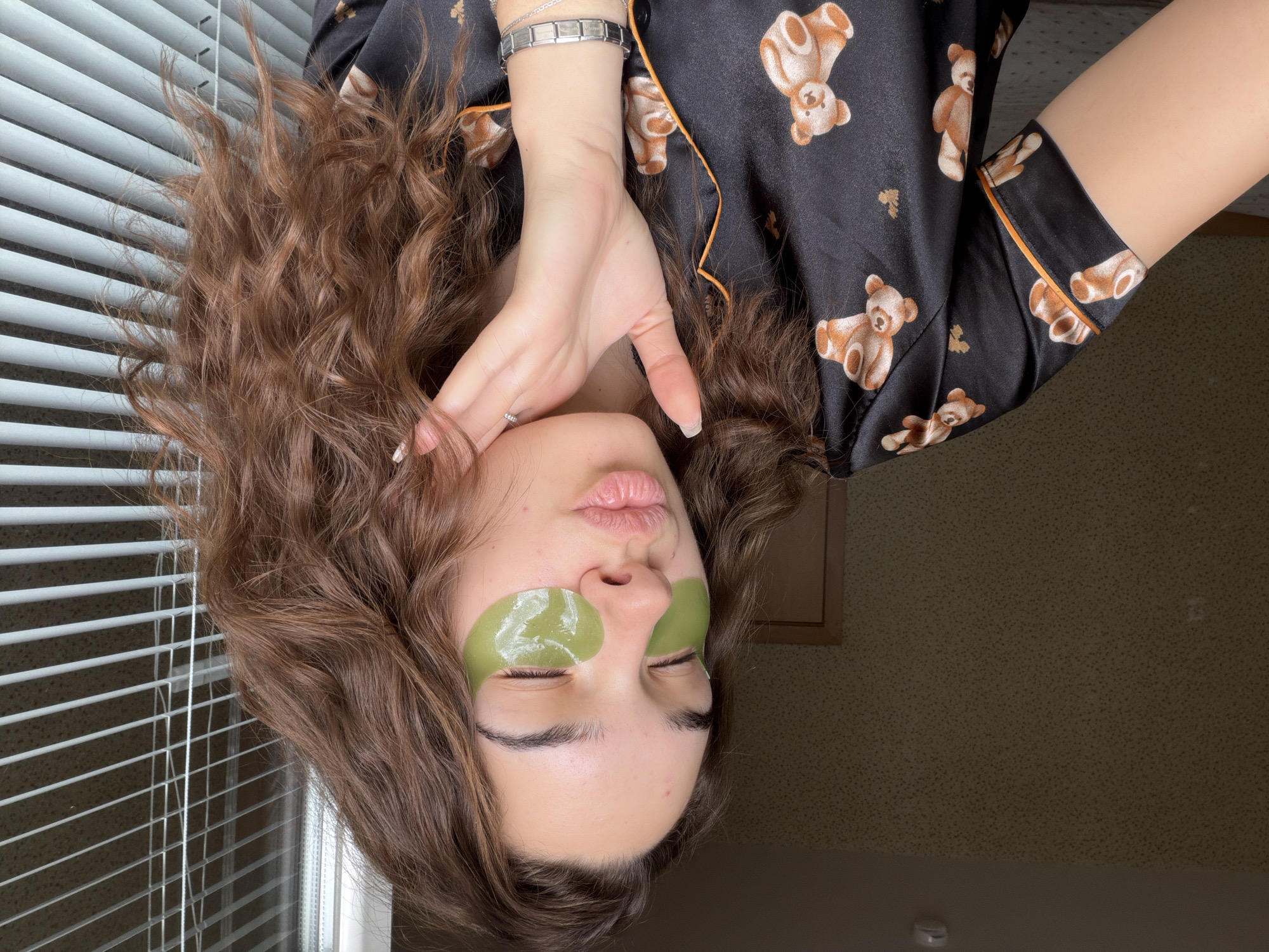 A young woman with long, curly brown hair is positioned upside down in the frame. She wears two green eye masks under her closed eyes and has her hand near her face. The woman wears a black pajama top with brown teddy bear designs and orange piping. Her left wrist sports a silver bracelet. Behind her is a set of white Venetian blinds and a beige wall with a patterned wallpaper. There is no information in the provided image that would suggest that the location is Sofia, Bulgaria.