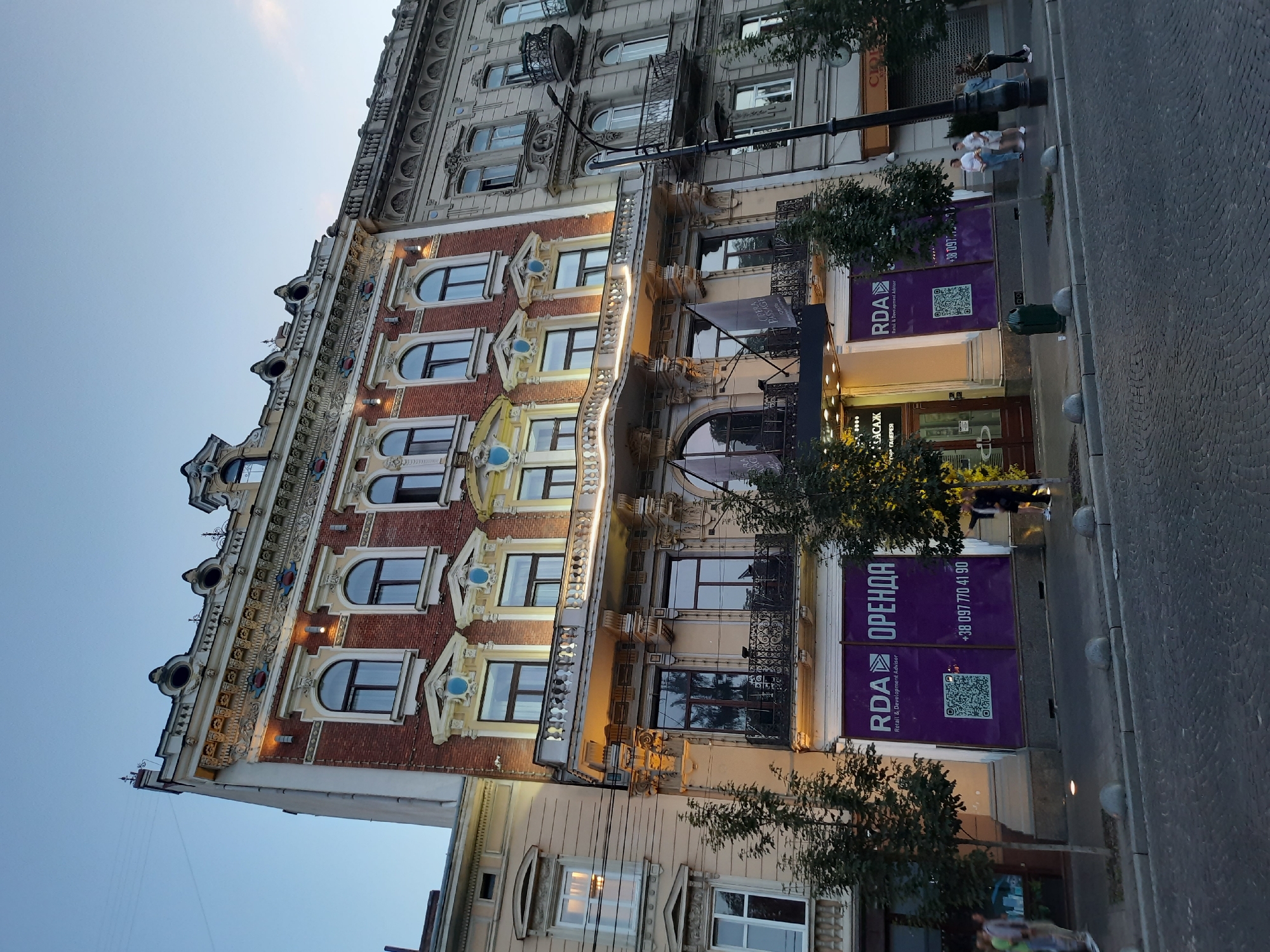 The image depicts a view of Rynok Square in Lviv, Ukraine, showcasing the architectural grandeur of the historic buildings. The main focus is on a multi-story building with an ornate brick facade, characterized by arched windows, intricate decorative elements, and balconies. The building is illuminated, highlighting its architectural details against the twilight sky.
In the foreground, a cobblestone street and sidewalk are visible, with a few trees planted along the sidewalk. Several people are walking on the sidewalk, and some are standing near the entrance of the building. One building in the scene has a large purple banner advertising "RDA ОРЕНДА" (RDA RENTAL) with a QR code and a phone number, indicating a commercial space for rent.
The adjacent building to the right is also historic, with a lighter-colored facade and similar architectural features like arched windows and balconies. A lamppost stands on the sidewalk, and a green trash can is present. The overall scene conveys a sense of urban heritage and commercial activity in a historic European city.