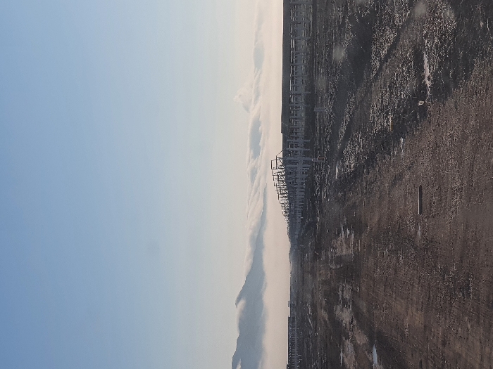 An expansive landscape unfolds under a gradient sky, transitioning from a clear blue to a pale, cloud-streaked horizon. Along the dividing line of sky and earth, a structure, possibly industrial, creates a repetitive, linear pattern. The ground is predominantly a rugged terrain of dark soil and debris, with subtle hints of moisture reflecting light. The overall scene presents a stark contrast between the natural sky and the human-altered land.