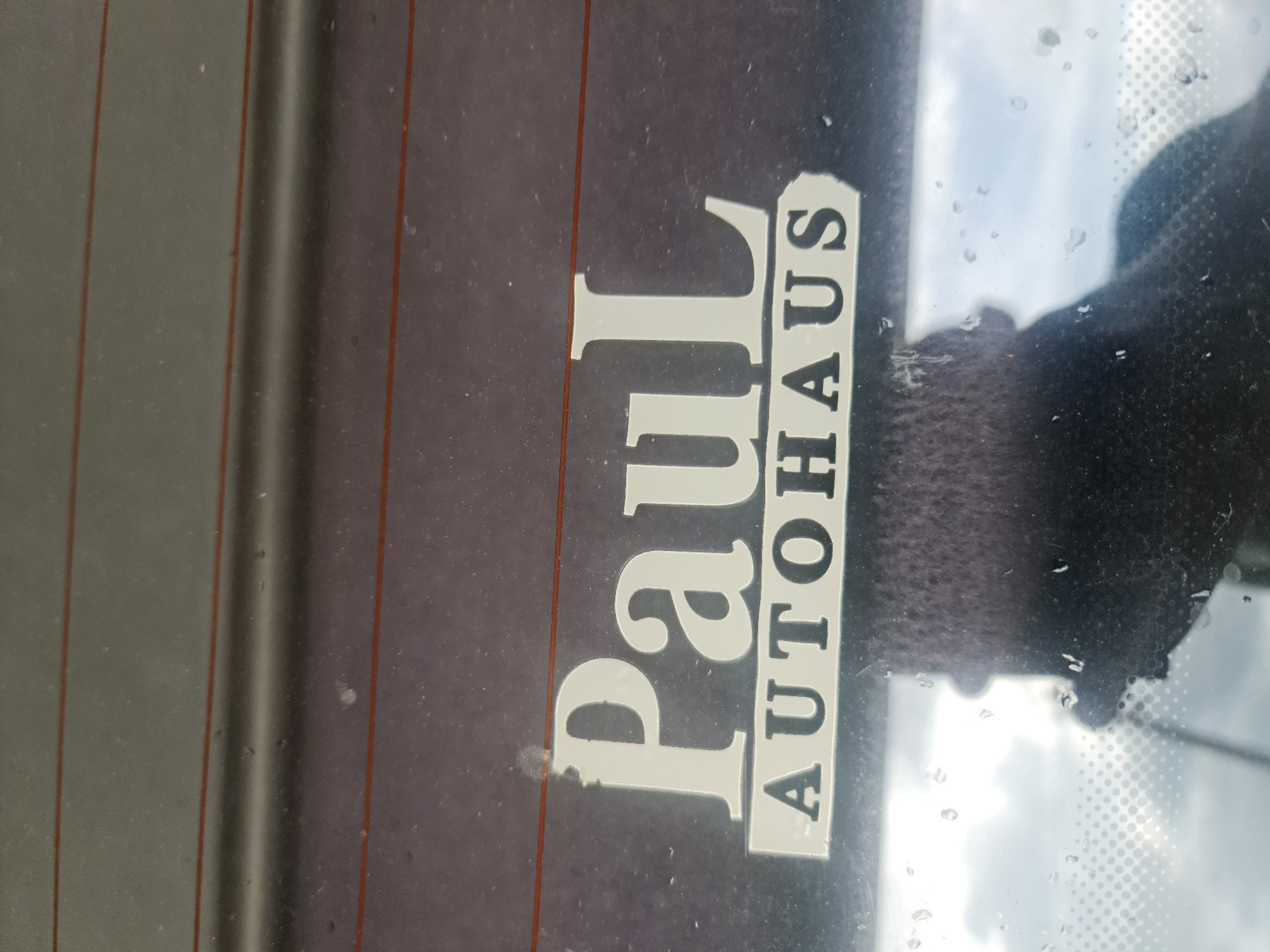 The image provides a close-up, slightly rotated view of a car's rear window, seen from the interior. Several reddish-brown horizontal lines, likely part of the window's defroster system, are visible across the surface. A prominent white sticker, partially worn or damaged towards the bottom left, is adhered to the window. The sticker displays the text "Paul AUTOHAUS." "Paul" is written in a large, bold, serif typeface, while "AUTOHAUS" is in smaller, sans-serif, uppercase letters, aligned vertically to the right of "Paul" and enclosed within a thin white rectangular outline.
The window is covered with numerous small water droplets, indicating either recent rain or condensation. Through the slightly obscured and wet glass, a bright, possibly cloudy sky is visible in the upper right quadrant of the image. A dark, textured object, possibly a car seat headrest or another interior element, is also partially discernible through the window. The overall brightness suggests it is daytime. The "AUTOHAUS" text on the sticker indicates a connection to a car dealership or automotive service, fitting within a general European context like Buzovgrad, Bulgaria.