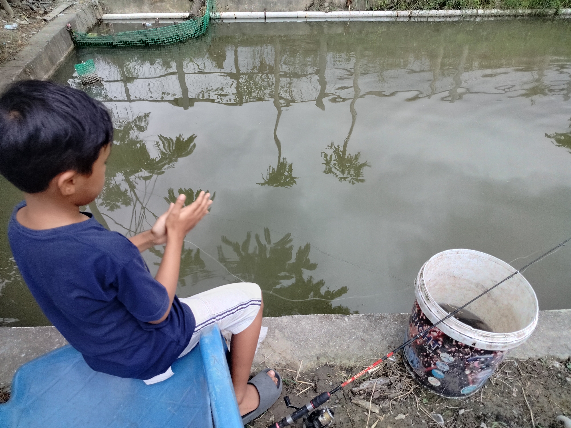 A young boy wearing a dark blue t-shirt and white shorts is sitting on a blue plastic chair by the edge of a pond. He is clapping his hands, facing away from the camera and towards the water. In the water, reflections of palm trees are visible. To the right of the boy, a white bucket with colorful designs sits on the ground next to a fishing rod. Inside the bucket, a fish is visible. The scene appears to be in Lembahalas, Indonesia.