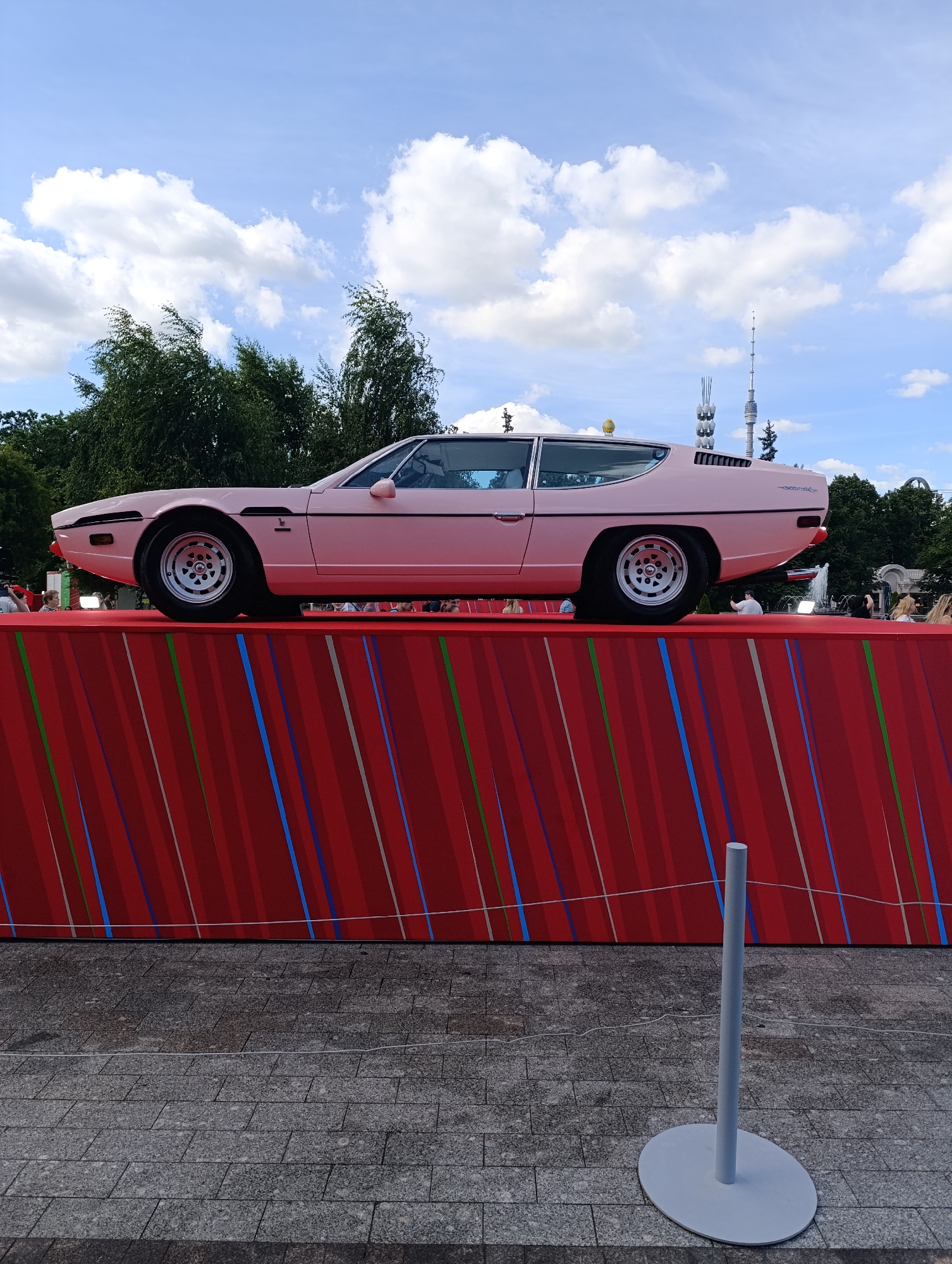 An eye-level, medium shot captures a vintage pink Lamborghini Espada parked on a red display platform in an outdoor urban setting. The car, viewed from its left side, is the central focus, with its sleek lines and classic design highlighted against a backdrop of a bright blue sky with scattered white clouds. Behind the car, a line of green trees forms a natural border, and several tall, distinct tower structures rise into the sky, including what appears to be a television tower. The platform on which the car rests is a vibrant red, adorned with a pattern of thin, diagonal stripes in various colors, including blue, green, and white. The foreground shows a grey, tiled ground with a simple, light-grey stanchion and its circular base standing to the right of the frame, with a thin white rope extending from it. In the background, to the left of the car and at the base of the platform, a few blurry figures are visible, suggesting the presence of onlookers. The overall scene is well-lit, indicating it was taken during daytime.