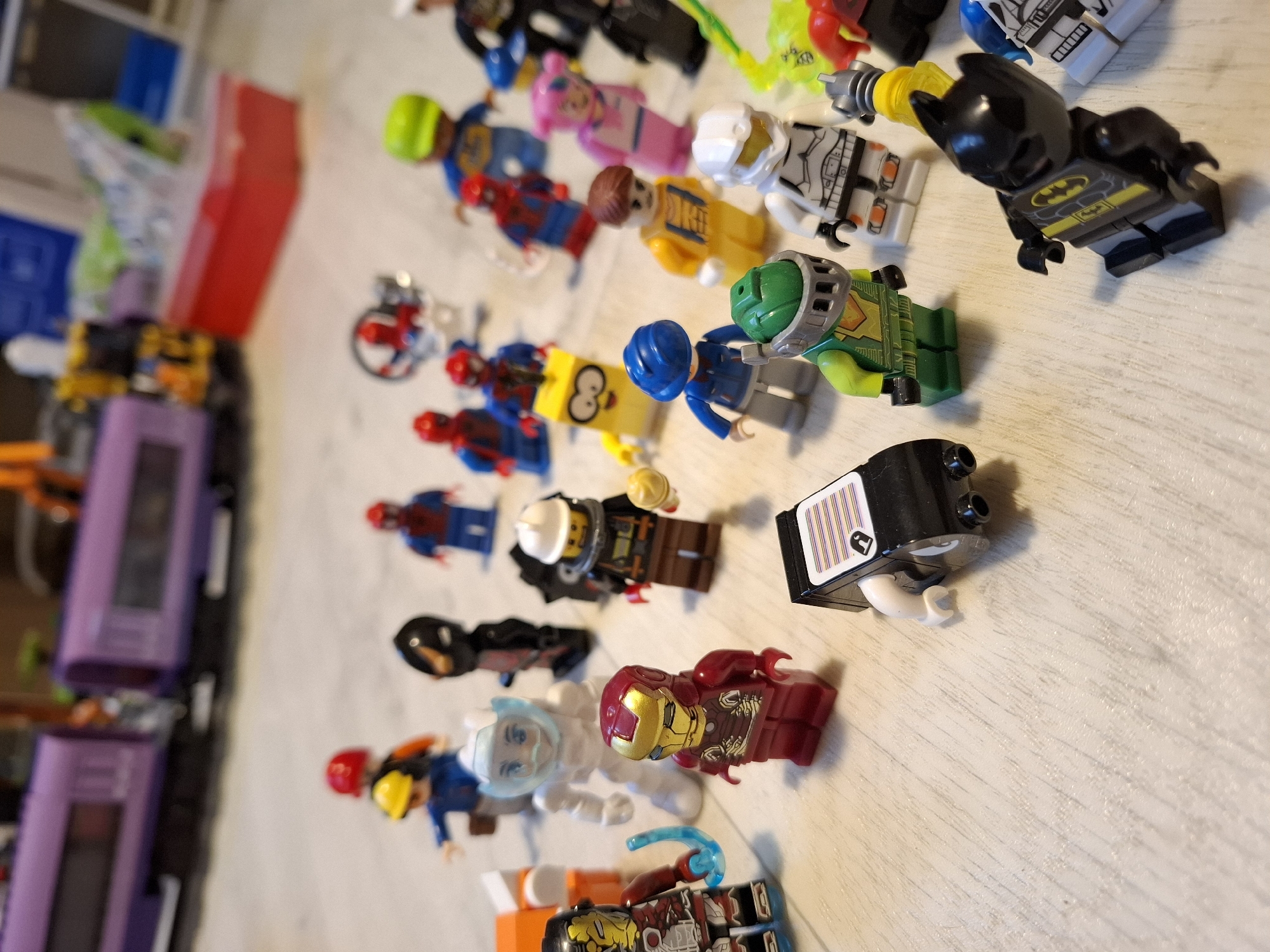 A collection of Lego minifigures are scattered on a light-colored, textured surface, which appears to be a wooden floor. In the background, a blurred purple Lego train is visible.
The minifigures are diverse, with various characters and costumes. Several appear to be superheroes, including at least one Iron Man figure, a Batman figure, and multiple Spider-Man figures. Other figures include a construction worker, a person in a white suit with a helmet, and a yellow character with large eyes.
The minifigures are positioned at different angles and in various poses, suggesting they have been placed or played with. Some figures are close together, while others are more spread out. There are no discernible interactions between the figures.
The lighting in the image is soft and appears to come from above, casting subtle shadows. The focus is on the minifigures in the foreground, with the background elements out of focus.
The image does not contain any elements that would indicate a specific location in the city of Tyumen, Russia. The scene is generic and could be anywhere with a flat surface and Lego toys.