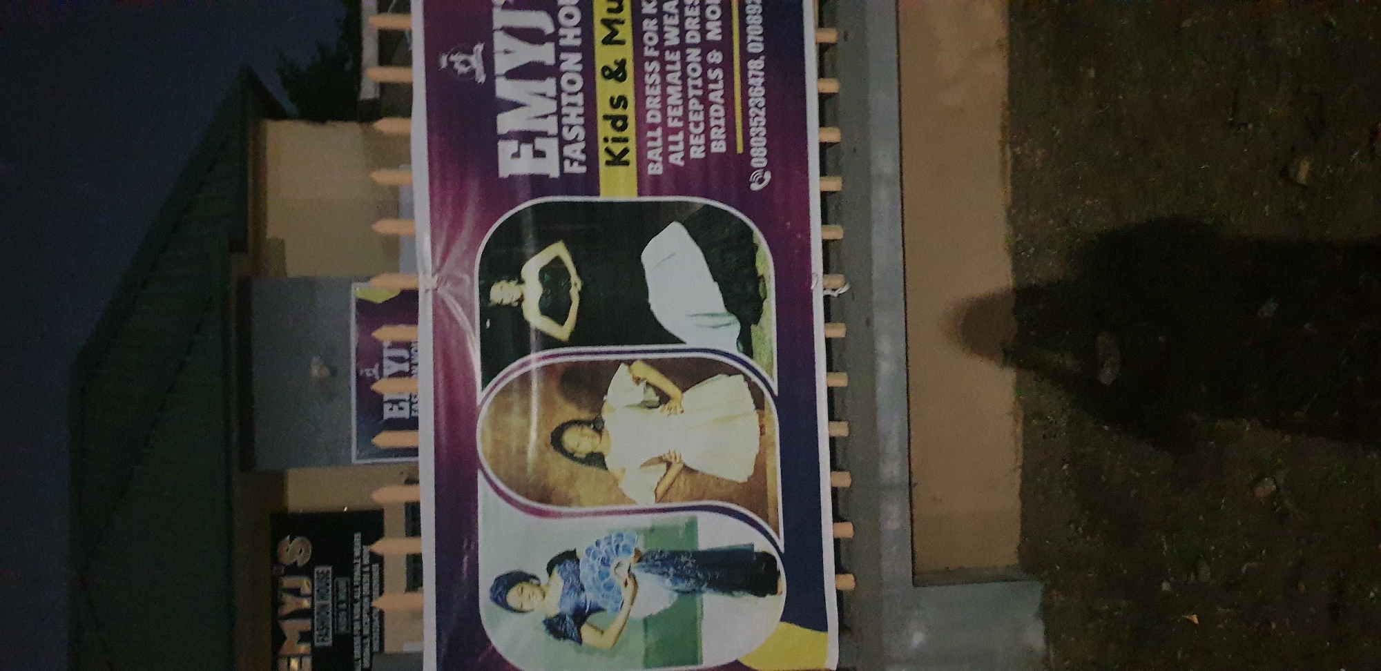 A banner advertising "EMYJ'S FASHION HOUSE" is displayed in front of a building with a green roof. The banner features images of three women in formal dresses and lists services such as "BALL DRESS FOR KIDS," "ALL FEMALE WEARS," "RECEPTION DRESS," and "BRIDALS." A phone number, "08035236478, 070892," is also visible. The scene appears to be in Narayi, Nigeria, based on the context. A shadow of a person is cast on the ground in the foreground. The lighting suggests it is evening or nighttime.