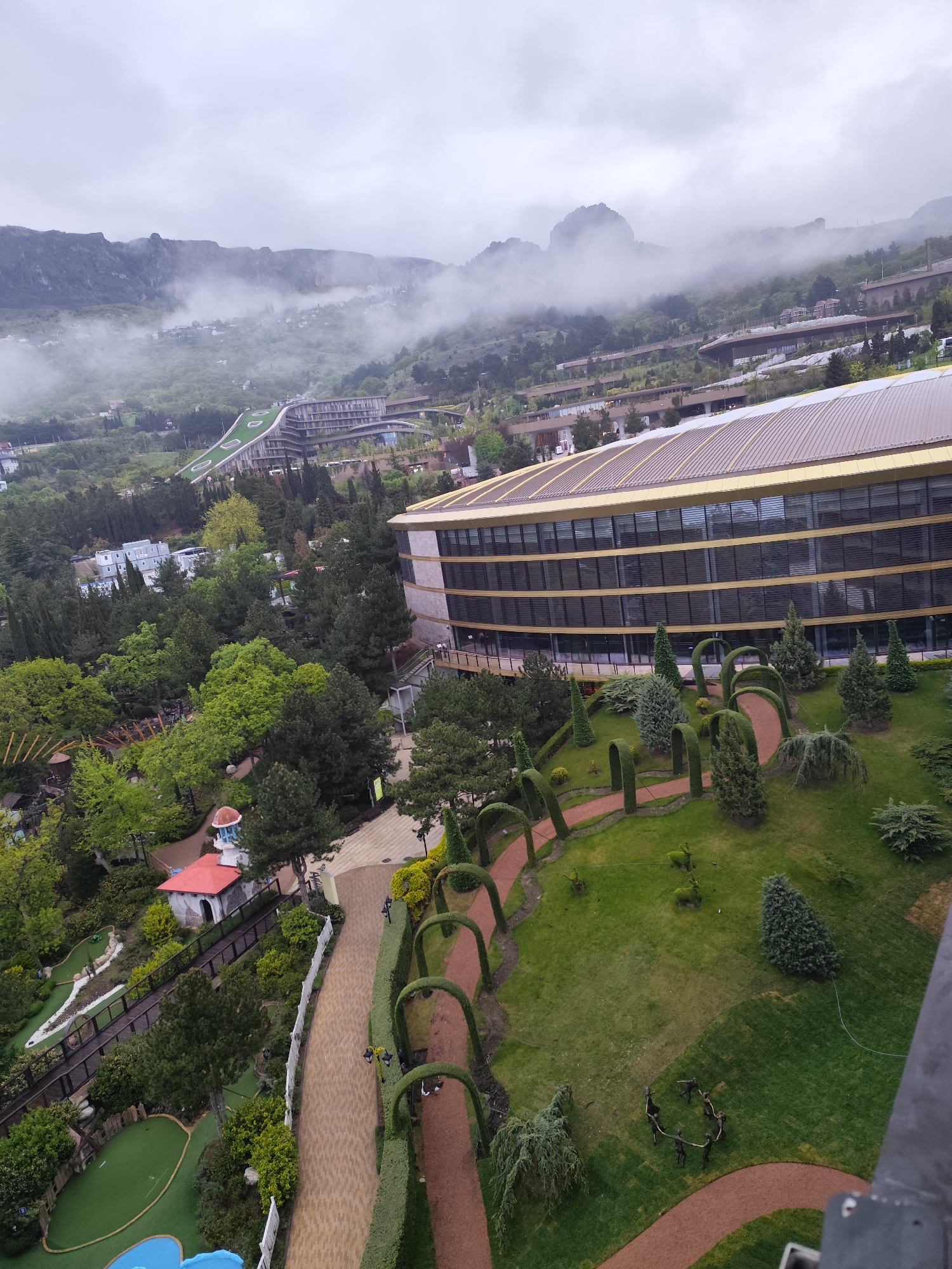 A high-angle view shows a portion of the city of Симеизский поселковый совет, Ukraine, on a cloudy day. Mountains are partially obscured by low-hanging clouds in the background. A multi-story, modern building with a gold-trimmed, curved roofline is a prominent feature in the midground. Adjacent to the building is a manicured garden with a series of arched hedges forming pathways. A mini-golf course is visible on the left, near a small building with a red roof and a blue turret. Paths wind through the landscape. In the distance, other buildings are nestled in the green hills.