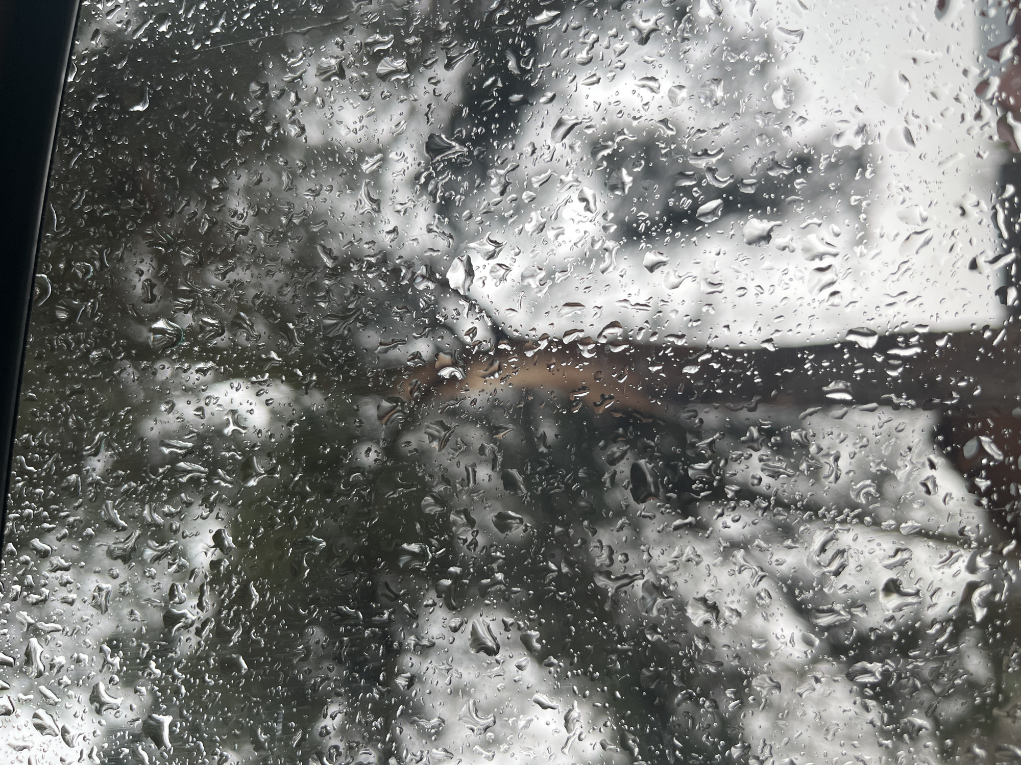 The image shows a close-up view of raindrops on a window. Through the blurred and distorted view, indistinct shapes and light suggest an outdoor scene, possibly a building and some vegetation. The location is unknown and cannot be verified to be the city of Razdolye, Russia. There are no people visible in the image. A portion of the black window frame is seen on the left side of the image. The primary subject is the raindrops obscuring the view of the outside.