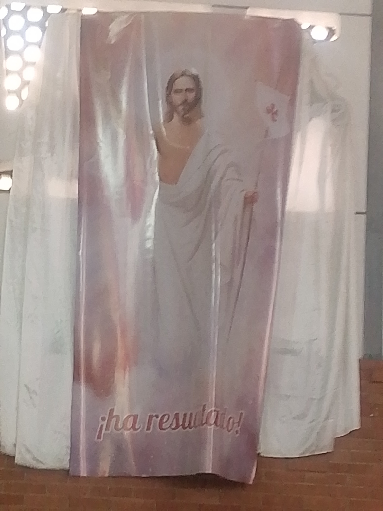 A rectangular banner featuring an image of Jesus Christ is centered in the frame. The banner displays the Spanish phrase "¡ha resucitado!" The banner is flanked by draped white fabric, possibly curtains. The floor appears to be brick or tile. The location might be Caracas, Venezuela, but that is not evident from the image itself. There is no evidence of people or interaction in the image.