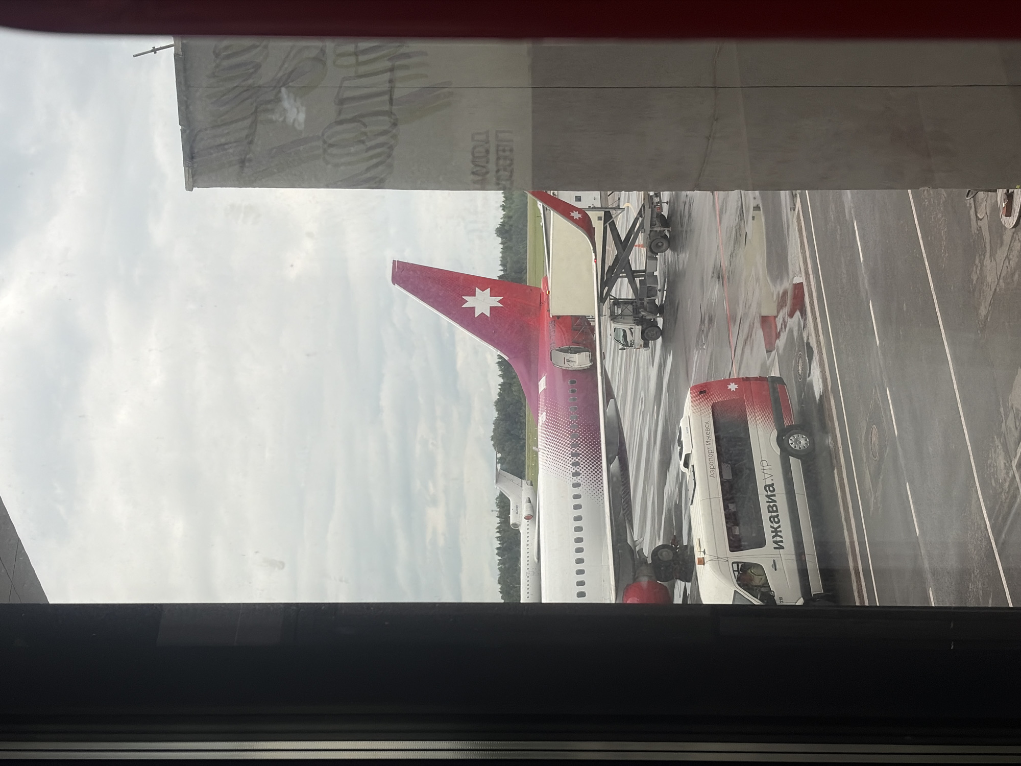 The image captures an airport tarmac scene viewed from an indoor window, with a dark frame in the foreground. The ground is wet, suggesting recent rain.
The central subject is the tail section of a passenger aircraft, oriented with its nose pointing right. The aircraft's vertical stabilizer (tail fin) is a distinctive red/maroon color, adorned with a prominent white, multi-pointed star emblem, characteristic of Izhavia airline. The fuselage is primarily white, transitioning to a subtle gradient or pattern of red/purple near the tail. Ground support equipment, including a baggage loader, is positioned next to the aircraft, indicating ongoing or impending ground operations.
To the right of the aircraft, a white and red airport shuttle bus is parked. Visible text on its side clearly reads "ИЖАВИА.VIP" and "Аэропорт Ижевск" (Izhevsk Airport), confirming the location as Izhevsk, Russia.
The sky is overcast, contributing to the muted lighting of the scene. The wet asphalt reflects the sky and the surrounding environment. Faint, mirrored text and patterns are visible as reflections on the window glass in the upper section of the image, partially obscuring the view of the sky and a building's roofline. The overall atmosphere depicts a typical daytime airport ground operation under cloudy weather conditions.