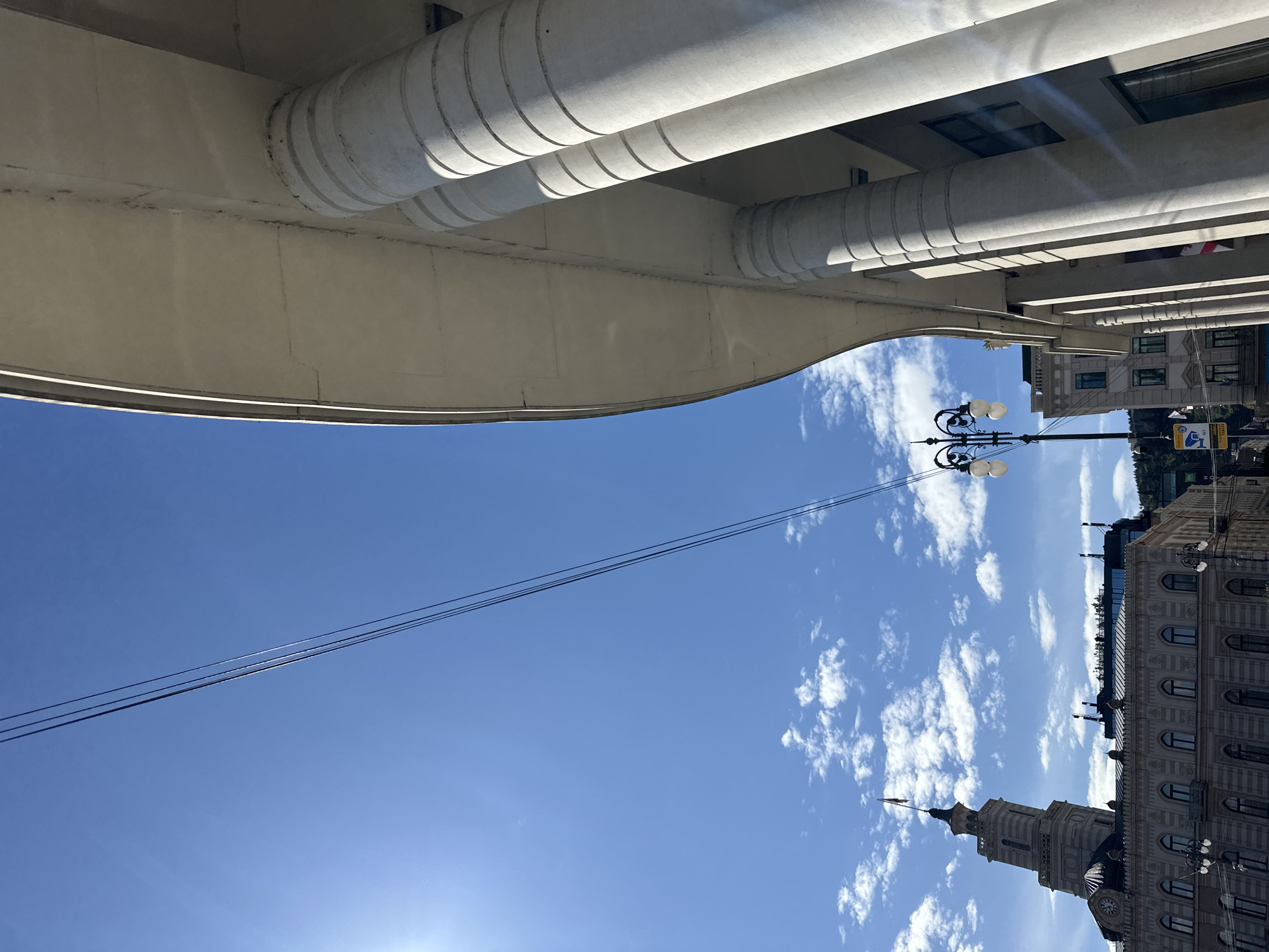 The image shows a street scene in Tbilisi, Georgia, taken from a low angle, looking up. Above is part of a bridge or building overhang, constructed with large, segmented, cylindrical supports. Below the bridge, the sky is blue with scattered white clouds. On the right side, buildings with classical architectural details are visible, including a prominent tower. A street lamp with multiple globe-shaped lights is also visible. Power lines stretch across the sky. A road sign is also visible. The image is oriented sideways.