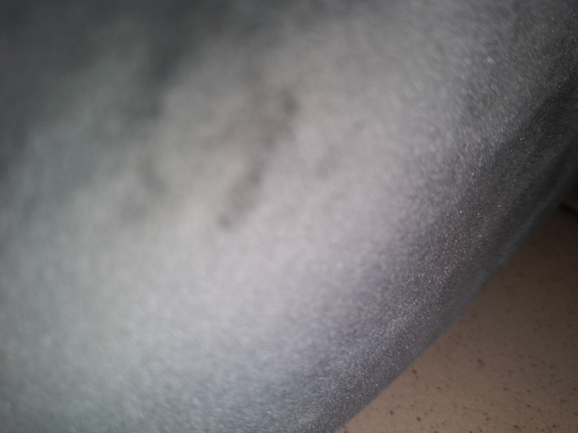 The image shows a close-up, out-of-focus view of a textured gray surface. The texture appears to be slightly grainy with some lighter, diffuse patches. A darker area occupies the right side of the frame, suggesting a shadow or a different material. The lighting is dim, contributing to the overall muted tones of the image. There are no discernible objects, people, or discernible environmental features that would allow for identification of a specific location within a city.