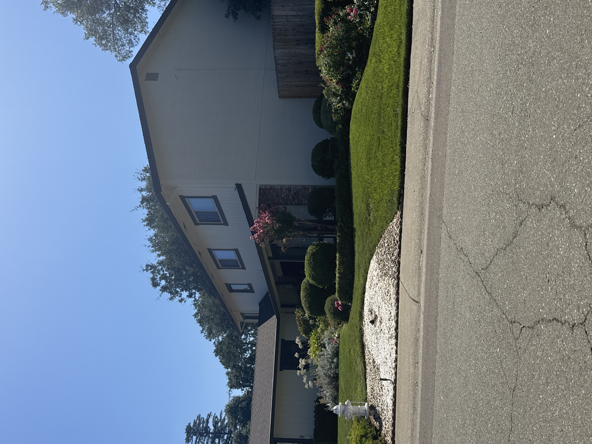The image depicts a suburban street scene in Citrus Heights, California, on a clear, sunny day. In the foreground, a cracked asphalt road runs horizontally, with a curb and a well-maintained lawn bordering it. A white fire hydrant is visible on the lawn, next to a circular bed of white gravel.
Behind the lawn, a two-story house with light-colored stucco siding and a dark-trimmed roof dominates the scene. The house has several windows, including a distinctive dark-trimmed horizontal window on the second story. A single-story section of the house, painted white with a dark-trimmed roof, extends to the left. This section has a dark-trimmed window.
The landscaping around the house is abundant, featuring manicured green bushes, a flowering pink tree, and a cluster of white flowers. A wooden fence runs along the right side of the property. Mature trees, including some oak trees, are visible in the background, and a clear blue sky stretches across the upper portion of the image. The scene suggests a residential area with well-kept properties.