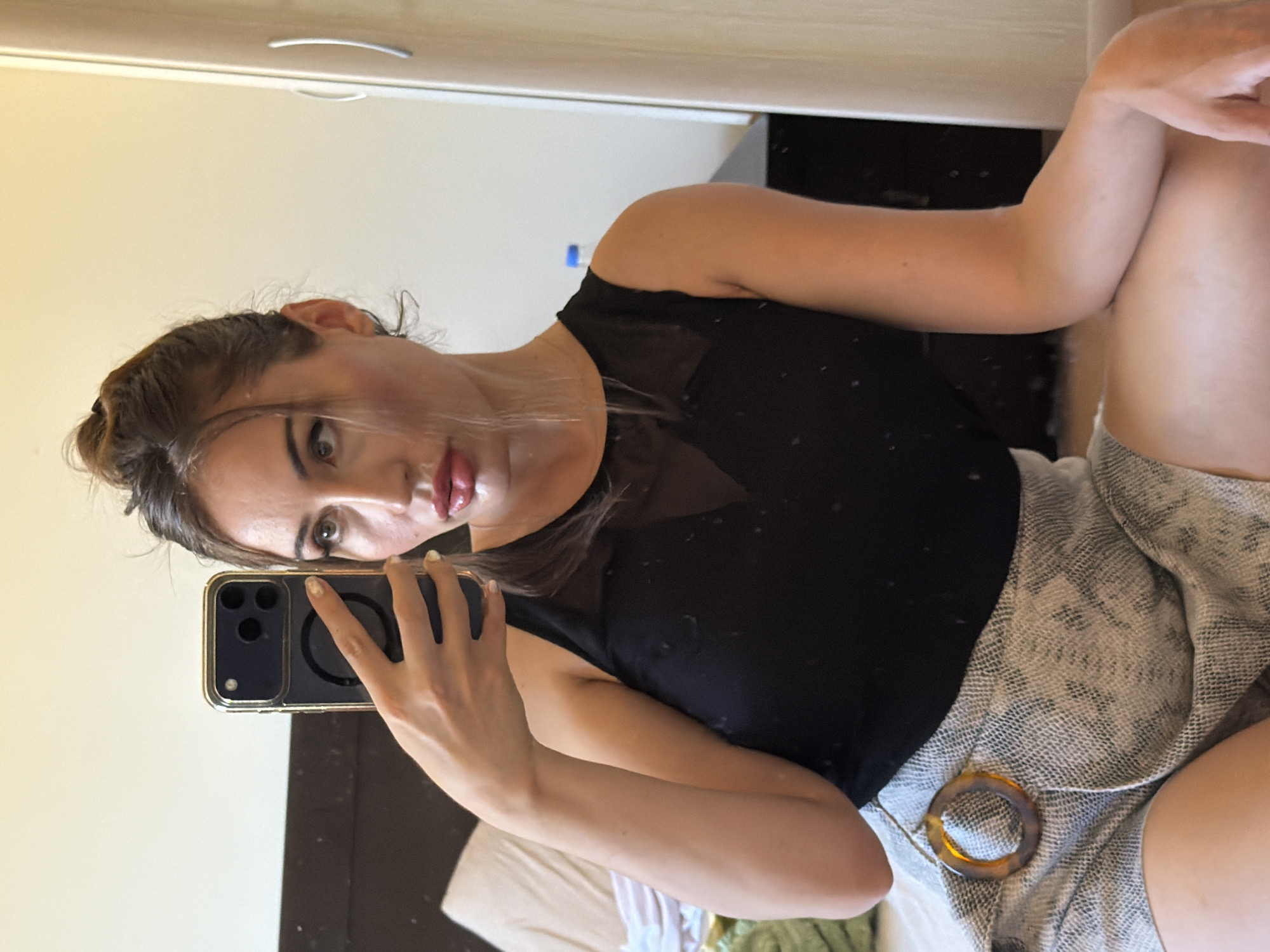 A woman is taking a selfie in a bedroom. She is holding a smartphone in her right hand, angled to capture her reflection in a mirror. The woman has dark hair pulled up in a bun and is wearing a black mesh-top tank and a textured grey skirt with a circular tortoiseshell-patterned buckle. She is sitting on the floor, with her legs crossed, in front of a bed. The room appears to be in the city of Tersefanou, Cyprus, based on the presence of a visible wardrobe typical of homes in the region. The lighting suggests it is daytime, possibly late afternoon.