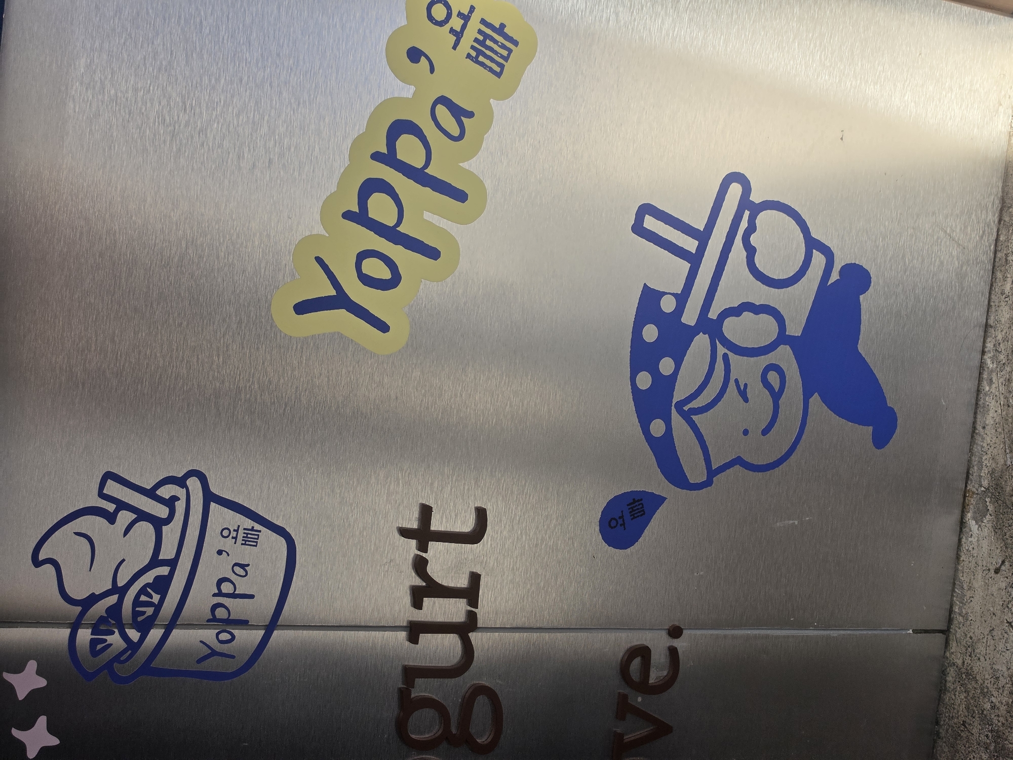 The image presents a close-up view of a brushed metallic surface, likely a sign or wall panel for a dessert or beverage shop in Bangkok, Thailand. The surface is adorned with several graphic stickers and partially visible text, predominantly in blue, yellow, and brown tones.
In the upper left, a bright yellow, cloud-shaped sticker features the brand name "Yoppa" in blue script. Adjacent to it, Korean characters read "여름" (yeoreum, meaning summer) and "요거트" (yogeoteu, meaning yogurt), translating to "Yoppa, Summer Yogurt."
On the right side of the image, a blue outline drawing depicts a stylized, cheerful character, possibly a girl, wearing a hat with dots and holding a cup with a straw, suggesting a drink like bubble tea. Below this character, the Korean word "여름" (summer) is visible in blue.
In the bottom left, another blue outline graphic shows a cup filled with soft-serve yogurt or ice cream, garnished with fruit slices (appearing to be lemons or limes) and a spoon or straw. Below this, "Yoppa" is written in blue script, followed by the Korean characters "여름" and "요거트."
At the very bottom of the frame, partially visible embossed-style brown letters spell out "gurt" and "ve.", hinting at a phrase such as "Yogurt Love." On the far left, fragments of small pink star shapes are also visible. The overall aesthetic is bright, playful, and refreshing, promoting a cool dessert or drink, likely during the summer season as indicated by the recurring Korean word for "summer."