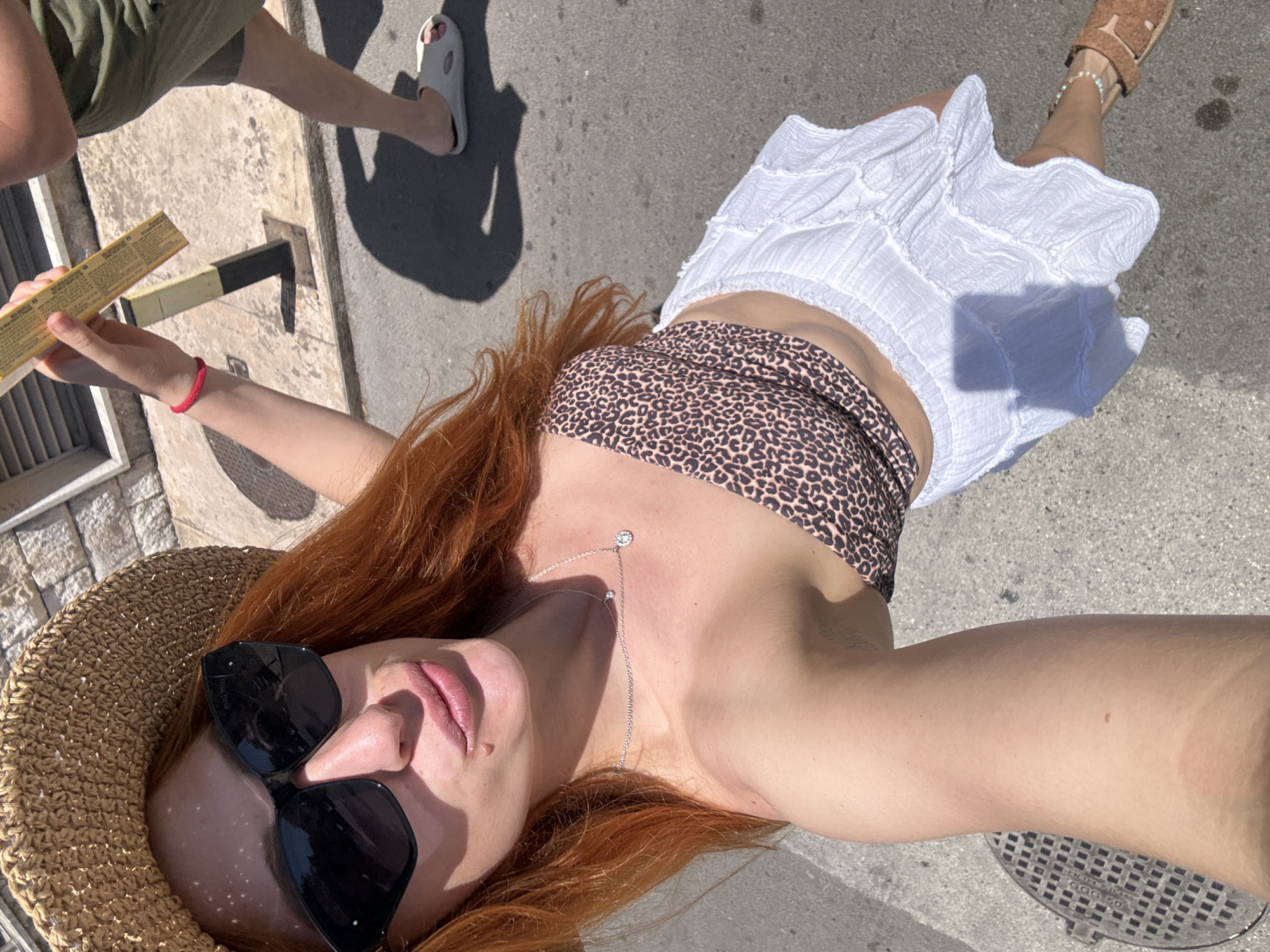 A woman with red hair, wearing a straw hat, sunglasses, a leopard print top, and a white skirt, is holding a rectangular object in her hand. She is standing on a grey pavement, presumably in a city. A leg wearing a green short and a white sandal is also visible, implying another person is nearby. The setting is Budva, Montenegro.