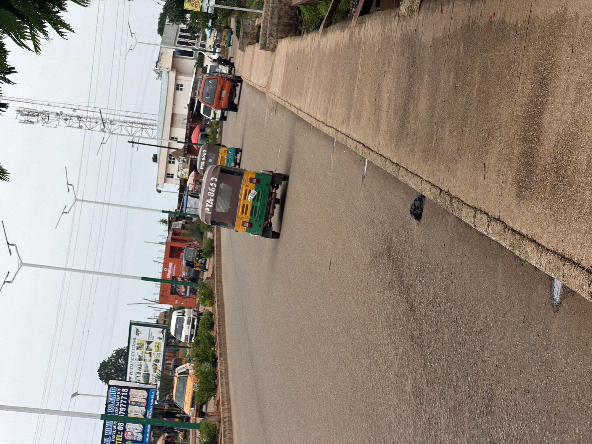 The image shows a street scene in Bukuru, Nigeria. A paved road dominates the foreground, with a concrete sidewalk running along the right side. Several vehicles are visible on the road, including a yellow taxi, a white van, an orange SUV, and three auto-rickshaws, also known as "keke" in Nigeria. One keke, with license plate PTA-8653, is in the foreground, facing away from the camera. Another keke, with license plate PTA-8087, is further up the road. Several people are visible, mostly near the vehicles and on the sidewalk, but their features are not discernible. Buildings and various signs are visible in the background, including advertisements and what appears to be a telecommunications tower. The sky is overcast. The overall scene suggests a busy urban environment with typical modes of transportation.