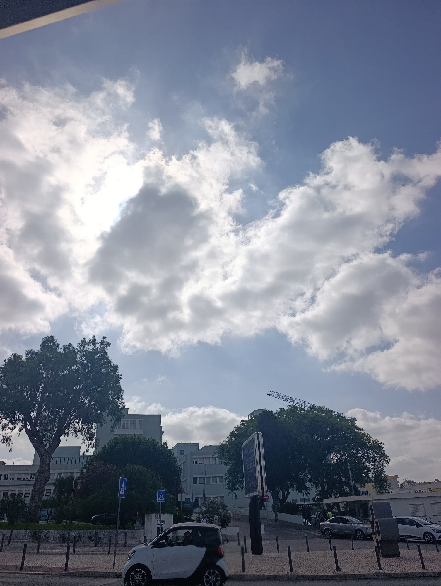A view of a modern building in Setúbal, Portugal, under a partly cloudy sky. The sun is bright and partially obscured by clouds. In the foreground, a white and black Smart car is parked on a cobblestone street. Across the street, there are several buildings, including what appears to be a hospital with a sign reading "HOSPITAL DE SÃO BERNARDO." There are trees lining the street and a construction crane is visible in the distance behind the trees. The scene depicts a typical urban environment on a bright day.