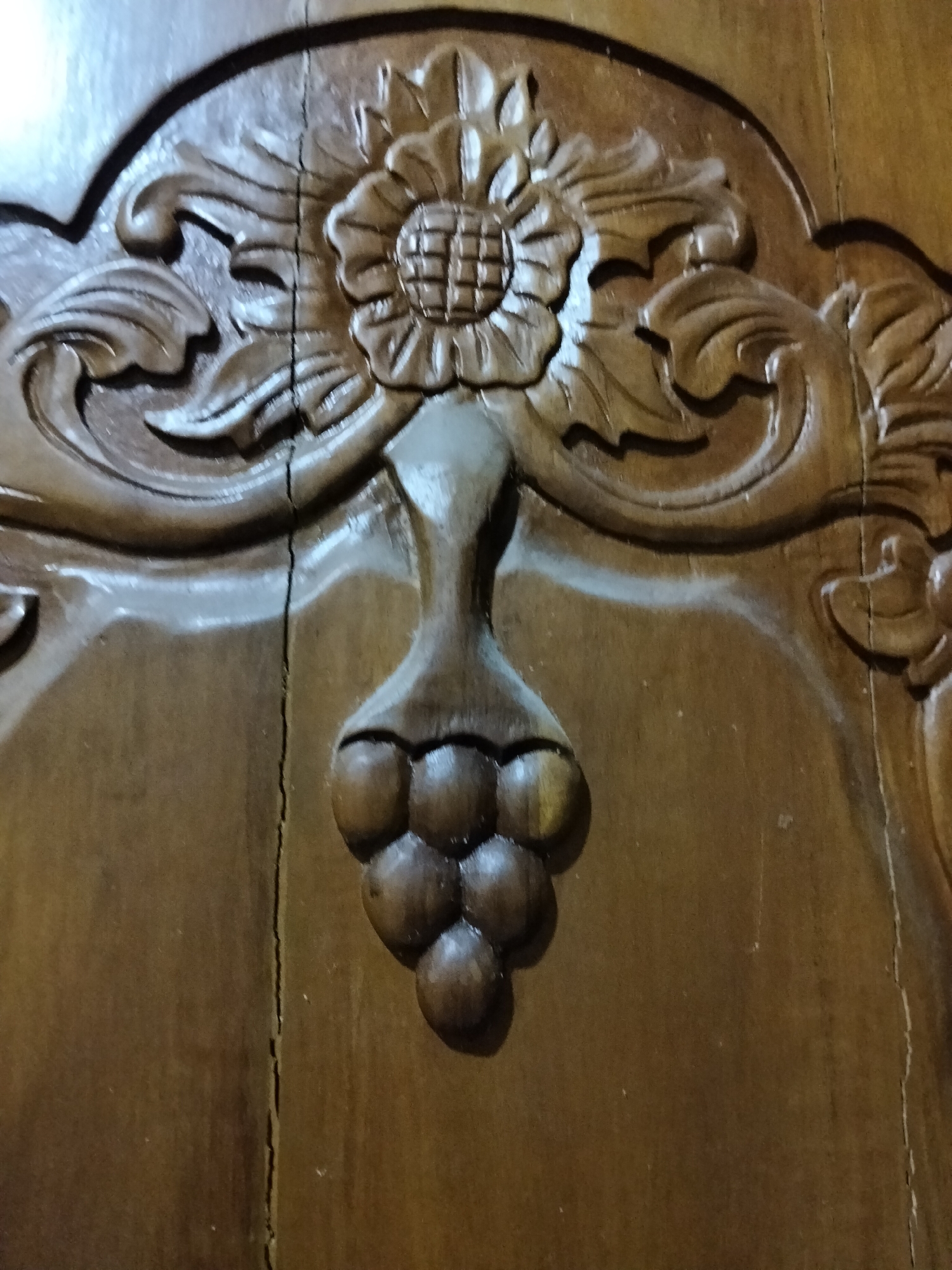 This media file shows a close-up of a wooden carving on what appears to be a piece of furniture or a decorative panel. The carving depicts a floral motif at the top, possibly a stylized sunflower, with ornate scrolling leaves extending outwards. Below the flower, a cluster of round shapes, resembling grapes, hangs from a stem-like element. The wood is dark brown and has a glossy finish, reflecting light in places. A prominent vertical crack runs down the center of the carved panel, disrupting the smoothness of the wood and the continuity of the design. The lighting suggests an indoor setting, possibly with artificial light casting shadows that highlight the depth and texture of the carving. The overall impression is one of traditional craftsmanship and decorative art. There is no visible text.