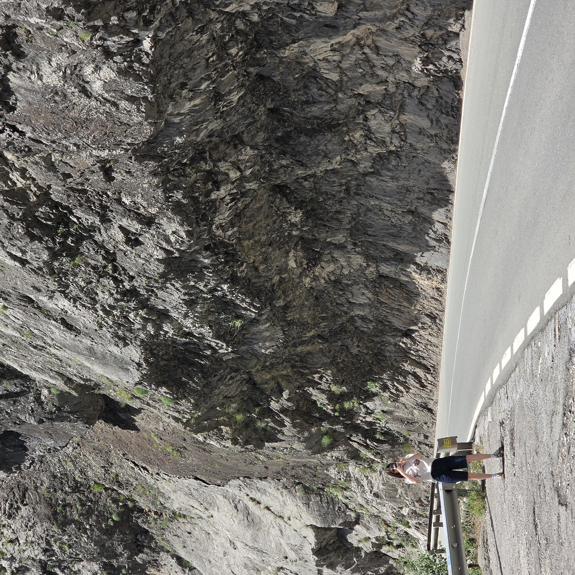 A medium shot shows a woman standing on a narrow roadside beside a steep, rocky cliff face. The woman is facing the camera and appears to be taking a selfie with her phone. She is wearing a short-sleeved white floral print shirt and dark blue shorts, with sunglasses resting on her head. She is standing next to a gray metal guardrail. The road surface is concrete and has a white center line. The cliff face to the left is composed of layered rock formations, some with sparse vegetation. The background is dominated by the gray and brown tones of the rock and the road. The location is not explicitly visible in the image, so the specification about Vlahovići, Montenegro, cannot be verified from the visual content alone.