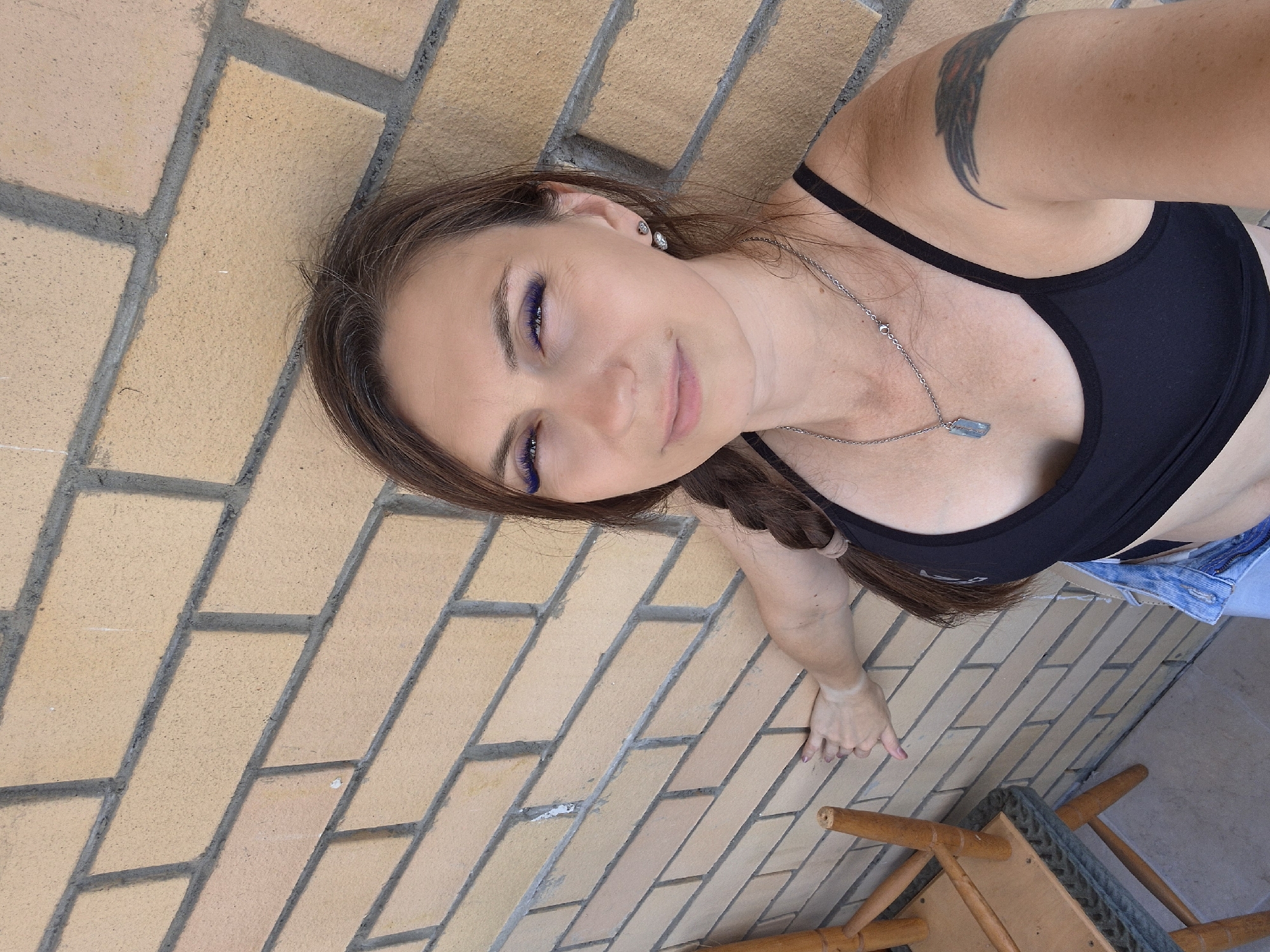 In a medium close-up, a woman with brown hair braided down her left side is positioned to the right of a wooden chair, leaning against a light brown brick wall. She is wearing a black sports bra, a silver necklace with a light blue pendant, and denim shorts. Her left arm is extended, with her hand resting on the brick wall. She has blue eyeshadow and long eyelashes. To the left of the woman, a section of a wooden chair with a woven seat is visible, its legs resting on a grey tiled floor. The background is an outdoor setting with a brick wall and a tiled floor, indicative of a balcony or patio. The scene is likely in Yekaterinburg, Russia, given the context.