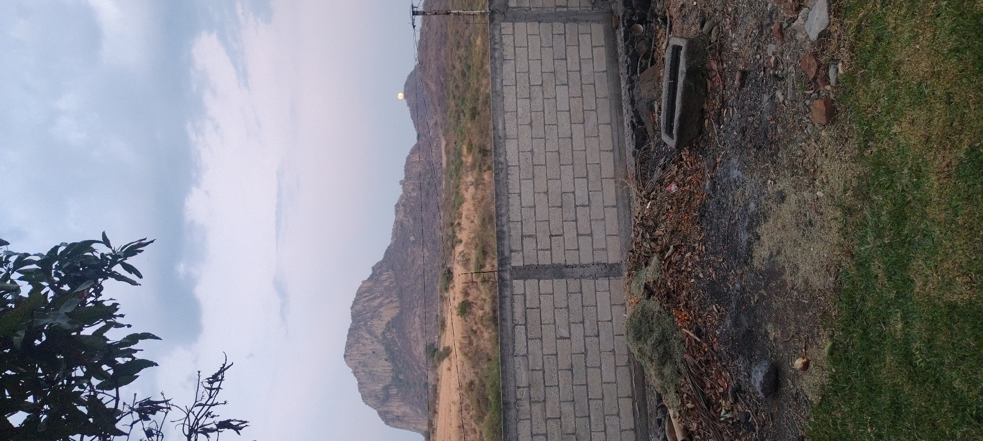 A landscape scene depicts a mountain range under a partly cloudy sky. A section of a brick wall extends along the right side of the frame, bordering an area of dry vegetation, bare earth, and a patch of green grass. In the foreground, a tree branch with green leaves partially obscures the left corner of the image. The sun is visible over the crest of the mountain range. Power lines run along the mountainside. The location is unknown.