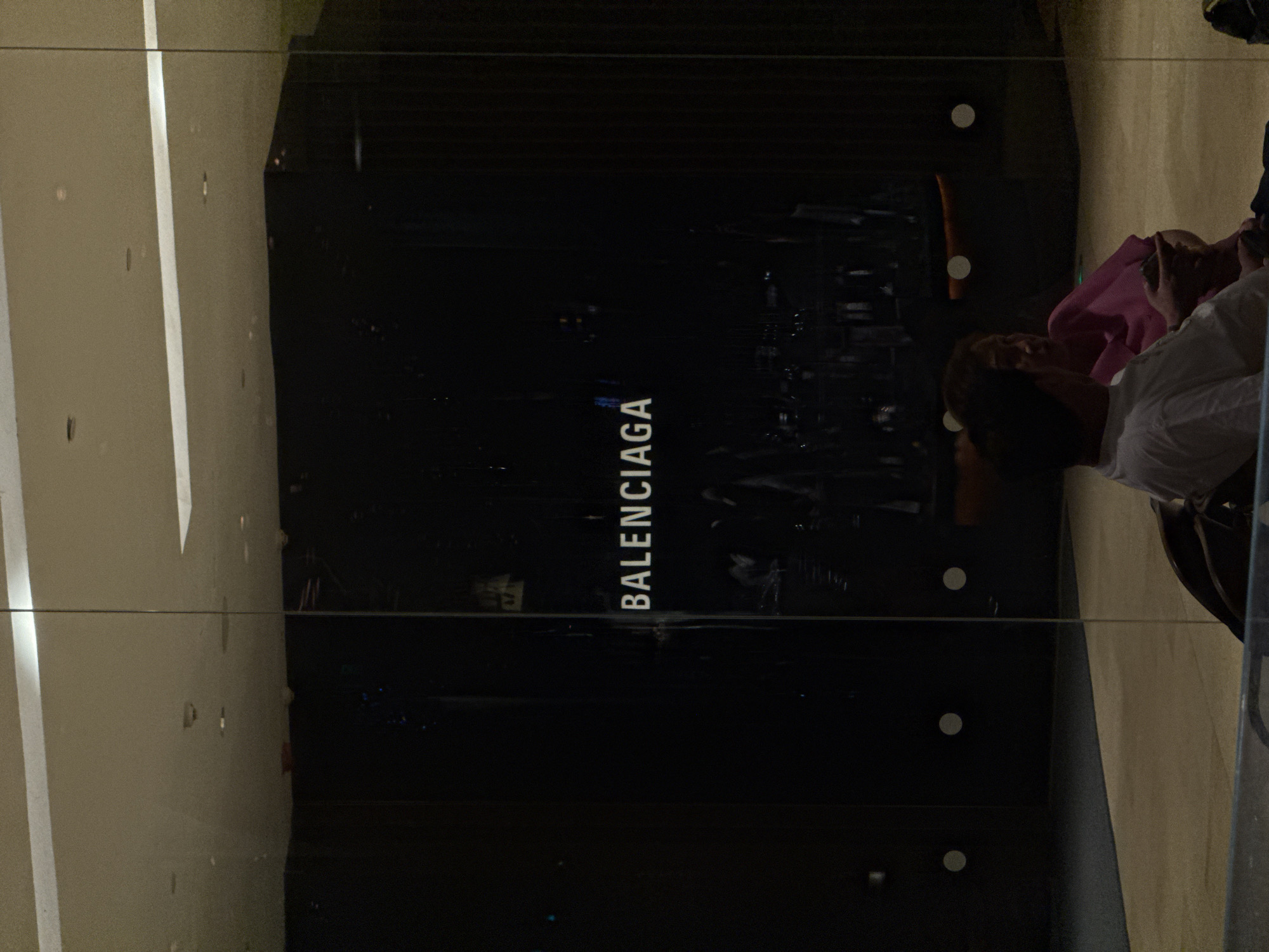 The image shows the front window display of a Balenciaga store. The store is located in Jing'an District, China. Two people can be seen reflected in the glass window. One is wearing a pink shirt and the other is wearing a white shirt. The store's signage, "Balenciaga", is vertically printed on the window. The background is dark, suggesting it is nighttime or the interior of the store is dimly lit. A horizontal wire is visible across the glass.